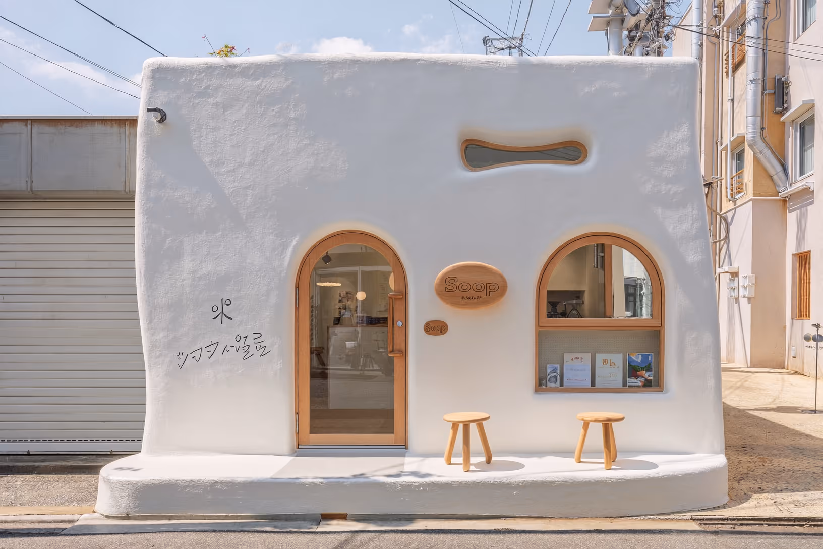 Petit bâtiment de plain-pied entièrement recouvert d'un enduit blanc lisse aux angles arrondis, avec deux ouvertures arquées en bois naturel et une fenêtre horizontale en forme de haricot, flanqué de deux tabourets en bois sur un trottoir urbain dense.