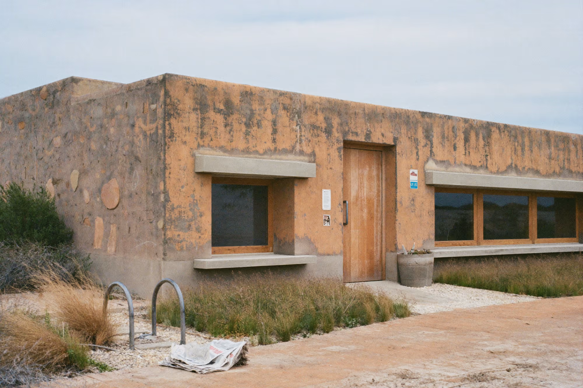 Façade en enduit ocre patiné avec inclusions de galets, porte en bois, fenêtres encadrées de béton, végétation sèche au premier plan, ciel couvert