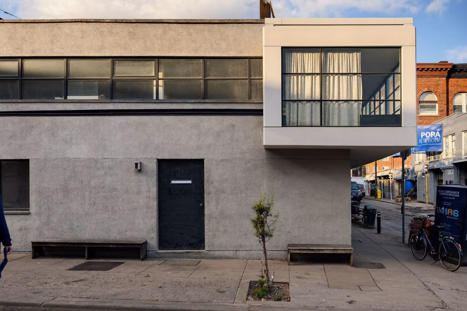 Façade urbaine composée d'un bâtiment bas en béton texturé gris avec porte noire, surmonté d'un volume blanc en porte-à-faux aux larges baies vitrées avec rideaux crème, dans une rue animée.