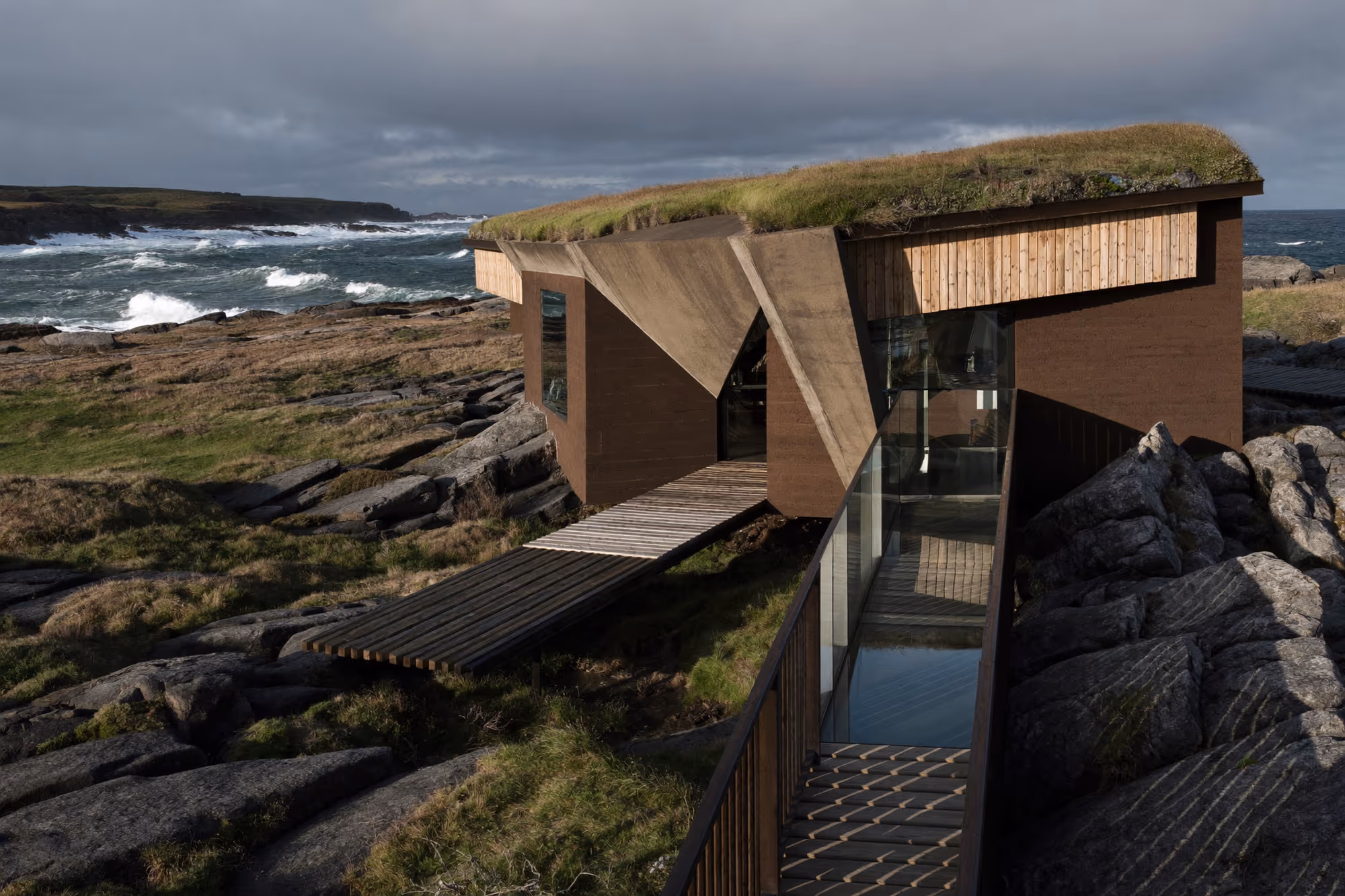 Deux volumes en bois brun foncé à toit végétalisé posés sur des rochers noirs au bord d'une mer agitée, reliés par une passerelle en bois, sous un ciel nuageux.