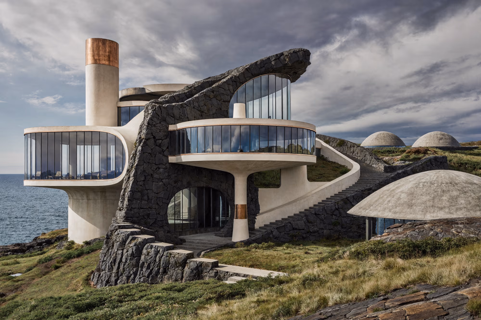 Bâtiment en béton aux volumes circulaires étagés sur un promontoire rocheux face à la mer, traversé par un affleurement de roche volcanique noire, avec des dômes semi-enterrés dans la prairie en arrière-plan.