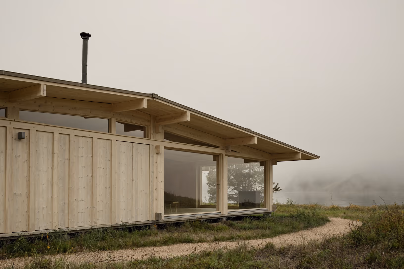 Maison basse en bois clair à structure poteau-poutre, toit débordant, grandes baies vitrées, entourée d'herbes hautes, sous un ciel brumeux et diffus.