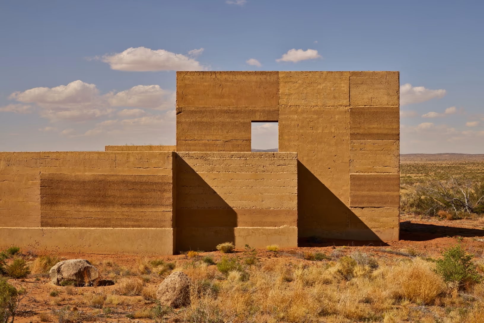 Structure en pisé aux strates horizontales marquées, posée dans un désert à sol rouge avec végétation rase, une seule ouverture rectangulaire découpée dans la masse, ciel bleu à nuages blancs en arrière-plan.