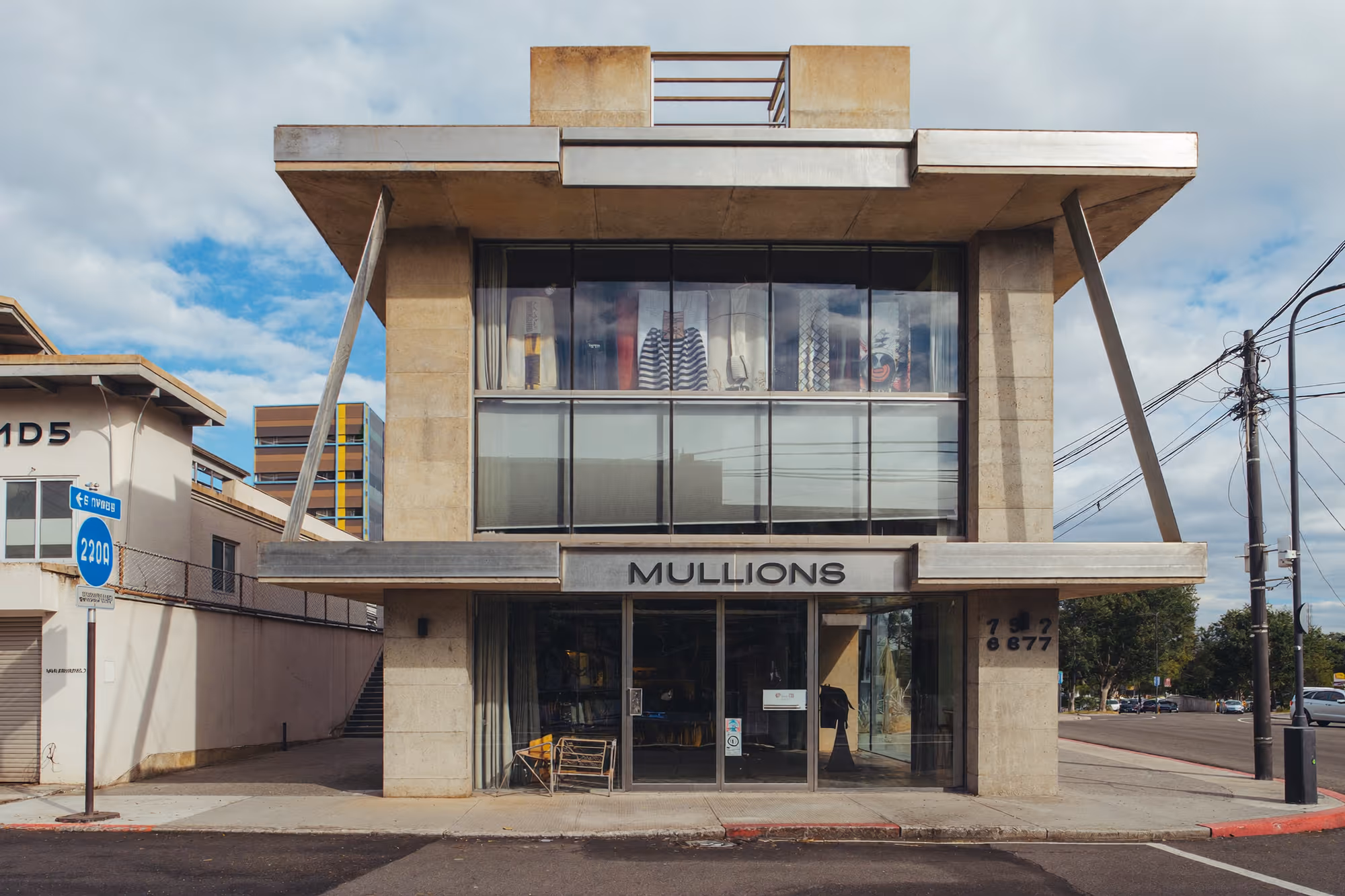 Bâtiment commercial de deux niveaux en béton brut, avec quatre piliers diagonaux inclinés vers l'extérieur, un bandeau vitré continu au premier étage, un toit en porte-à-faux avec garde-corps métallique, et une entrée vitrée au rez-de-chaussée, photographié en vue frontale depuis la rue.