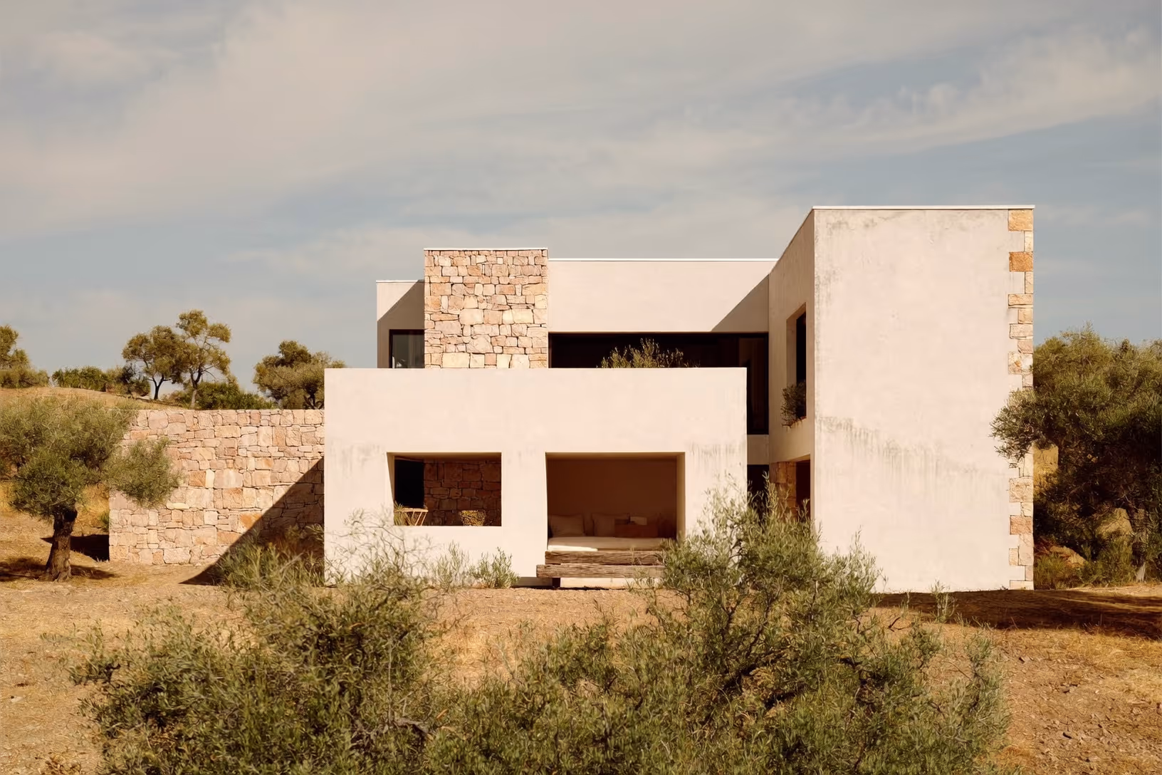 Maison cubique à deux niveaux, volumes blancs enduits contrastant avec des parements en pierre calcaire dorée, entourée d'oliviers et d'arbustes de garrigue sur un terrain sec et pierreux.