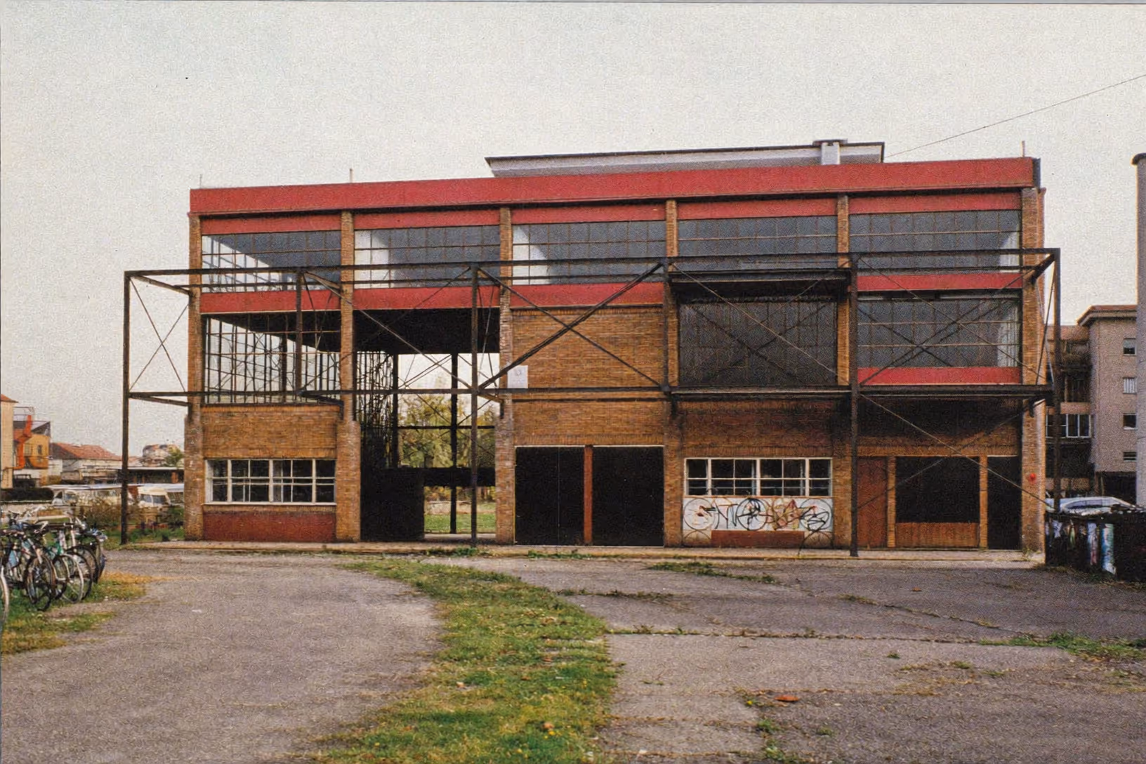 Bâtiment industriel à deux niveaux : rez-de-chaussée en brique jaune avec portes et fenêtres basses, étage vitré encadré d'une ossature métallique à contreventements en croix, bandeau rouge sous la toiture plate. Parvis asphalté enherbé, ciel couvert, vélos garés à gauche, graffiti sur les murs du bas.