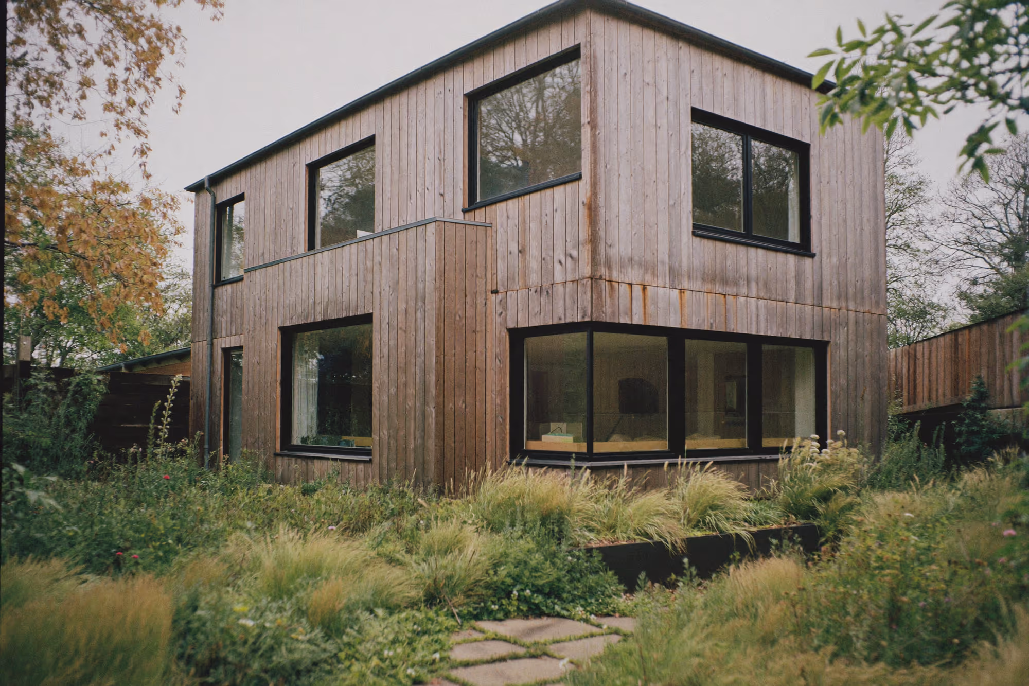 Maison à deux niveaux avec bardage bois vertical gris-brun, menuiseries noires, grandes baies vitrées au rez-de-chaussée, entourée d'une végétation dense et naturalisée sous un ciel couvert.