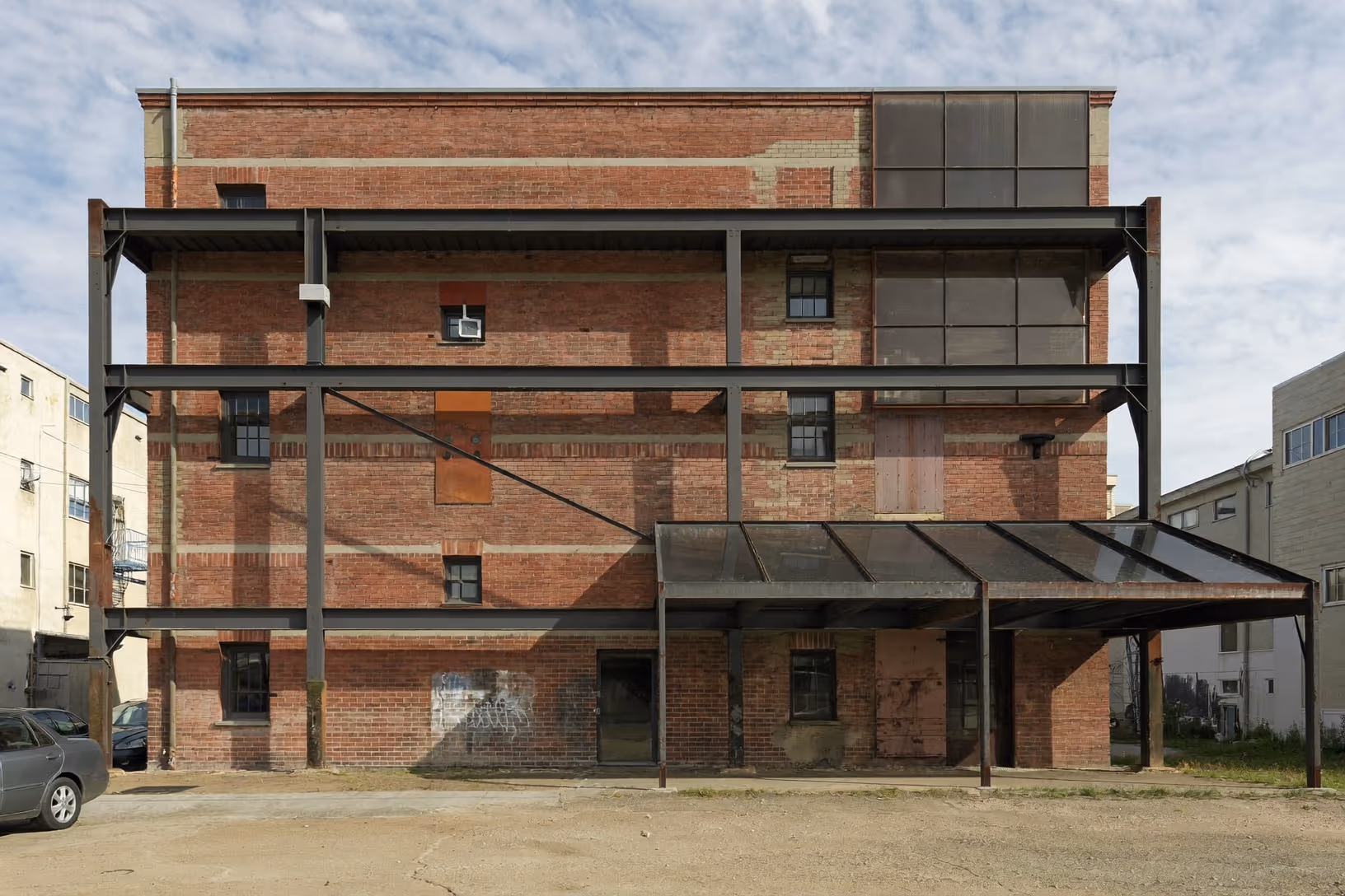 Façade d'un bâtiment industriel en brique rouge de quatre niveaux, avec une structure secondaire en profilés d'acier sombre apposée en avant-corps, une marquise inclinée en métal au rez-de-chaussée, fenêtres à petits carreaux, graffitis au bas du mur, dans un contexte urbain dense sous ciel nuageux.