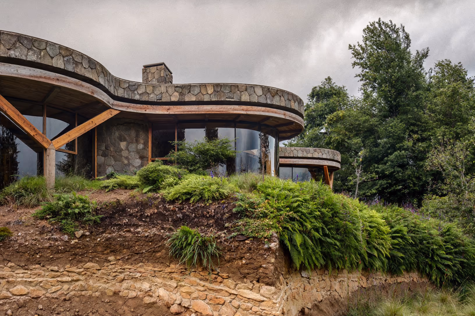 Maison aux contours arrondis construite en pierre et bois sur un talus végétalisé, entourée de fougères et d'arbres feuillus sous un ciel nuageux.