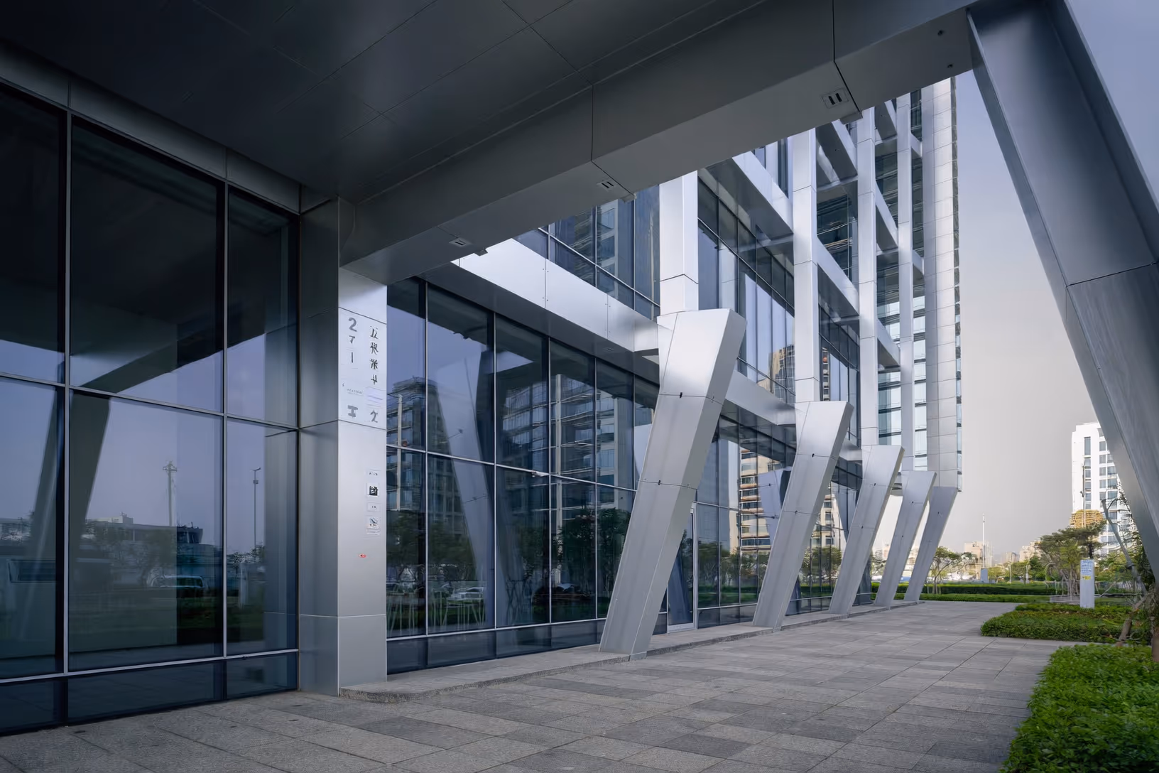 Façade d'un bâtiment contemporain composée de grandes baies vitrées sombres et de piliers diagonaux en métal blanc laqué, sous un auvent horizontal en acier gris, avec dallage en granit et végétation taillée au premier plan.
