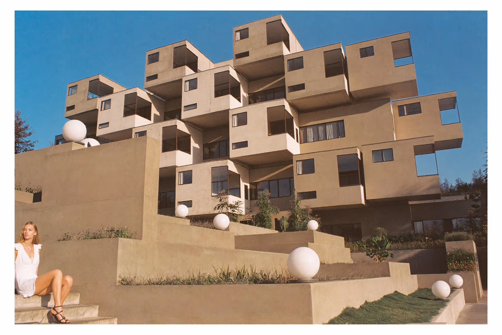 Ensemble résidentiel en béton enduit beige composé de volumes cubiques empilés et décalés, avec terrasses en gradins et sphères lumineuses blanches au premier plan, sous un ciel bleu dégagé.