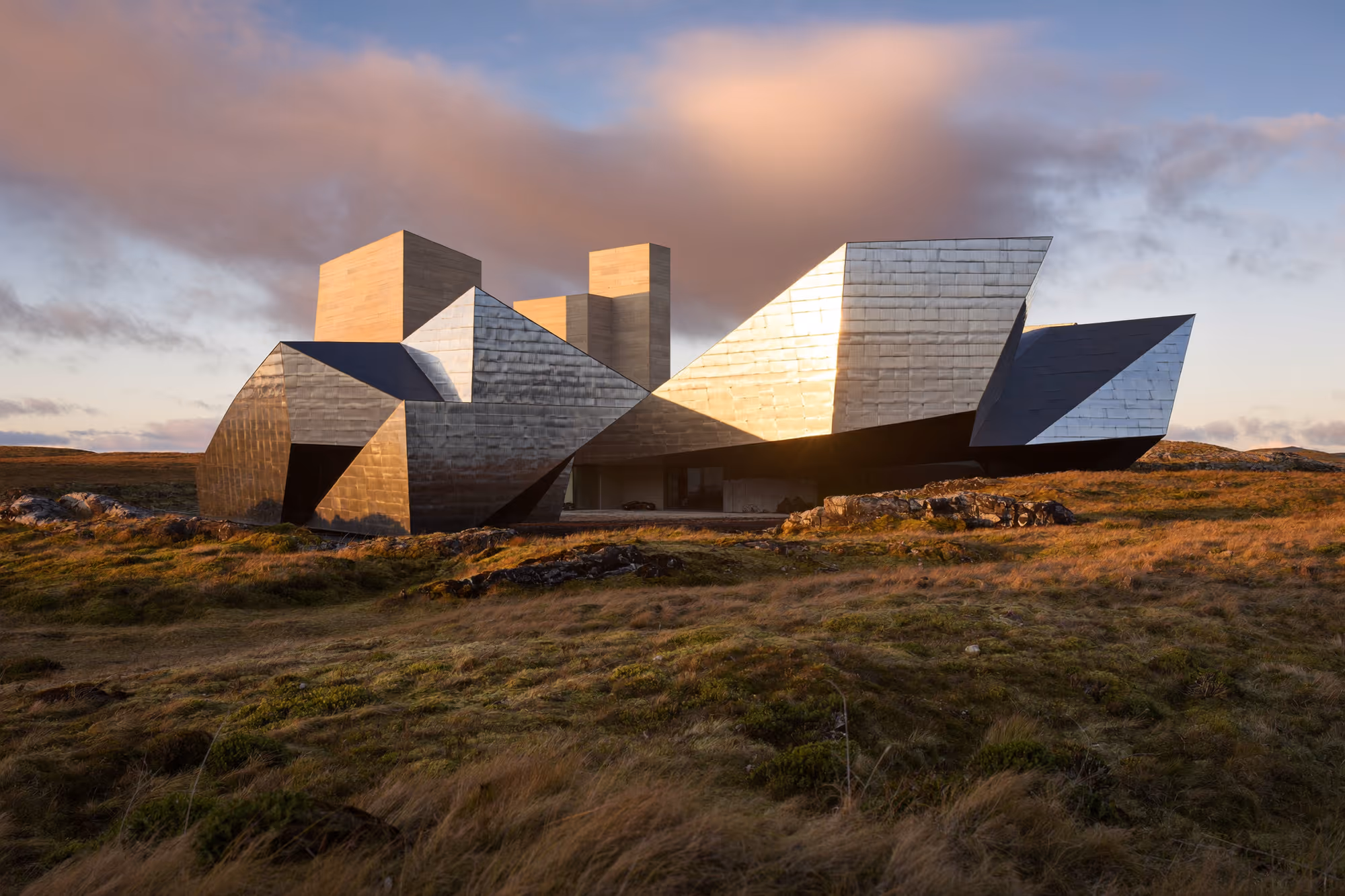 Structure architecturale aux volumes géométriques multifacettes recouverts d'ardoise gris argenté, posée sur une lande rase et ocre sous un ciel de crépuscule aux nuages rosés
