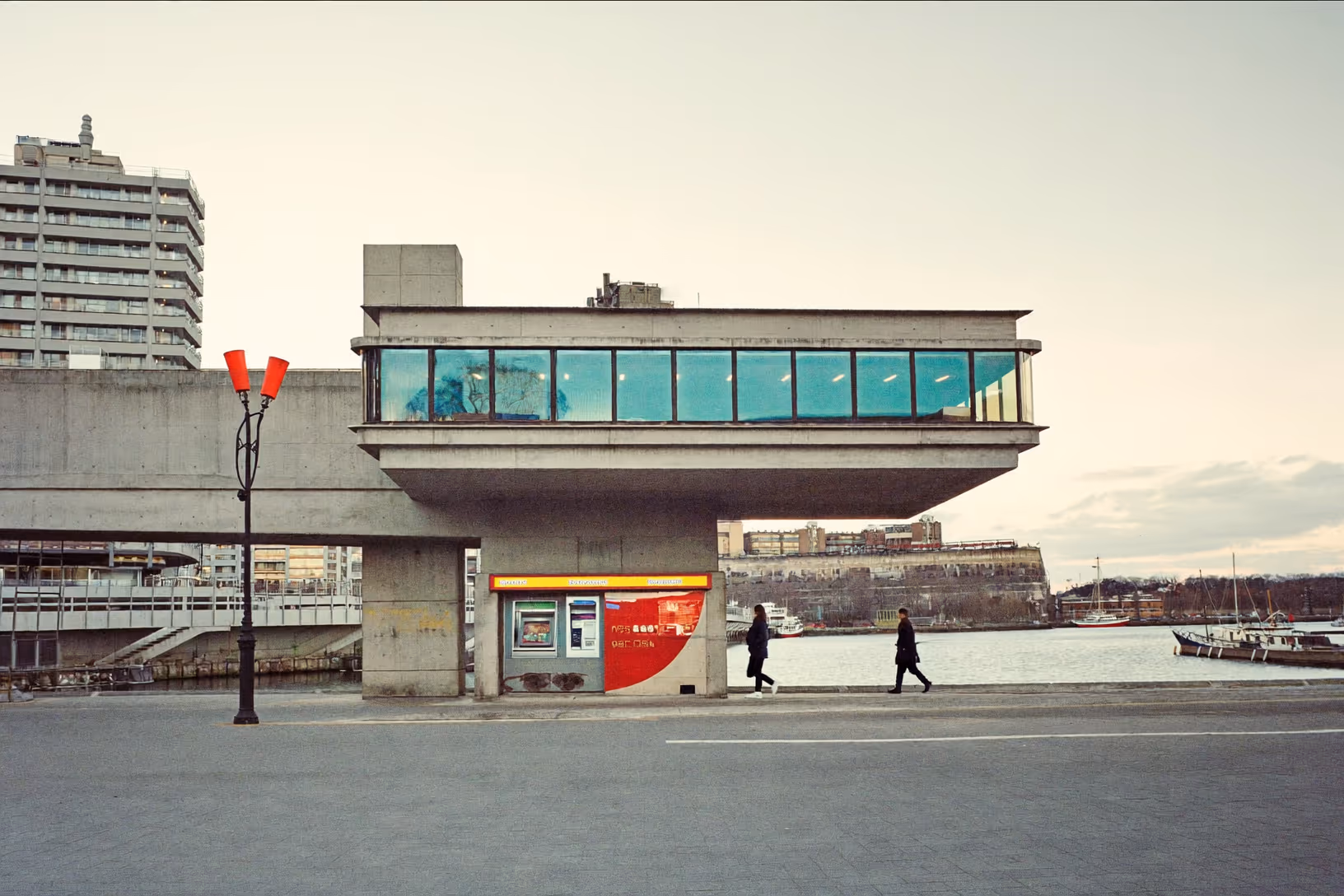 Édifice brutaliste en béton avec un étage vitré en porte-à-faux, situé sur un quai au bord de l'eau en hiver, avec des passants et un kiosque rouge au rez-de-chaussée.