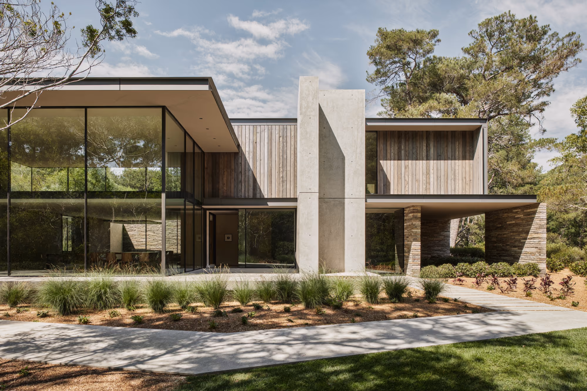 Façade d'une maison contemporaine à deux niveaux dans un sous-bois de pins, avec un pilier central en béton brut, un bardage bois en lames verticales claires, un soubassement en pierre appareillée et de larges baies vitrées à chassis noirs.