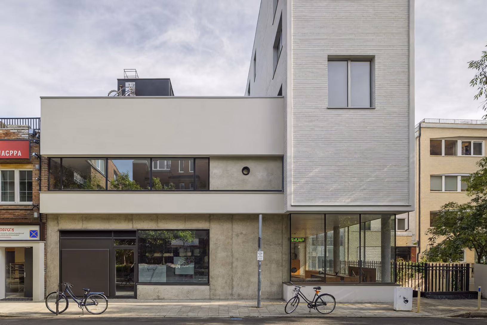 Façade d'angle d'un bâtiment contemporain en milieu urbain dense : rez-de-chaussée en béton brut avec larges vitrages, volume intermédiaire en enduit clair percé d'une fenêtre bandeau, tour étroite en brique claire avec fenêtre carrée, vélos au premier plan.