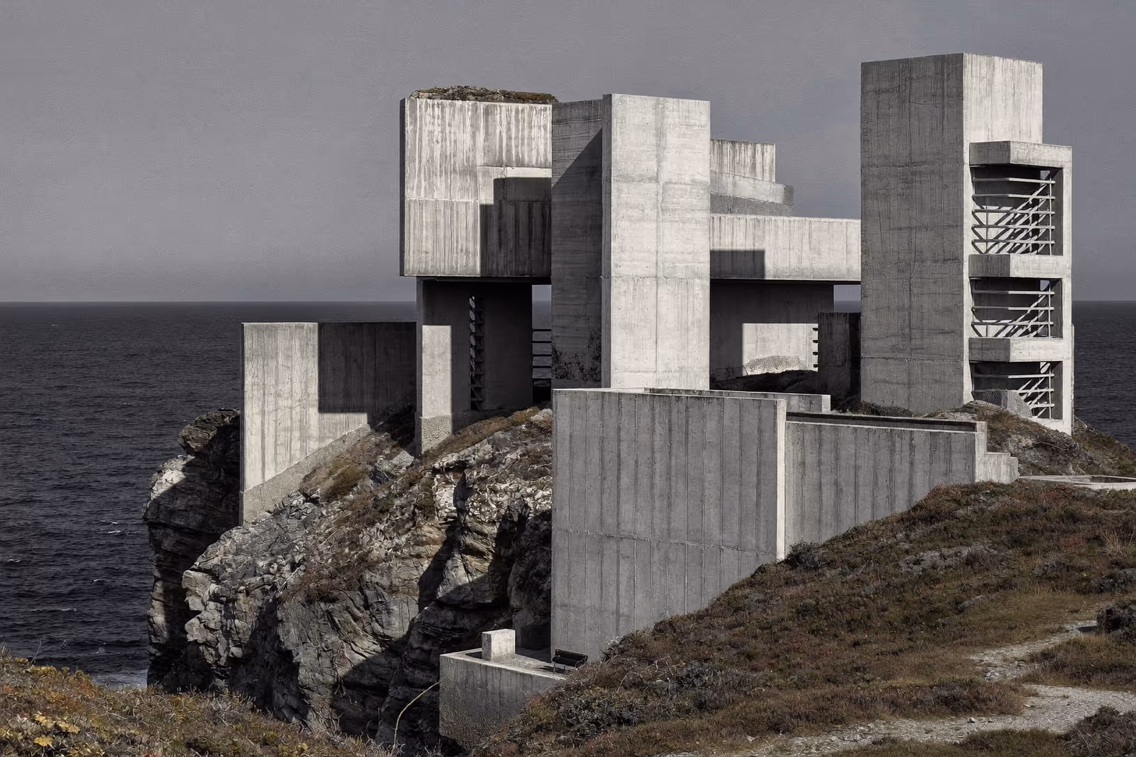 Structure monumentale en béton brut composée de volumes cubiques en porte-à-faux, posée sur une falaise rocheuse au bord d'une mer grise, sous un ciel couvert.