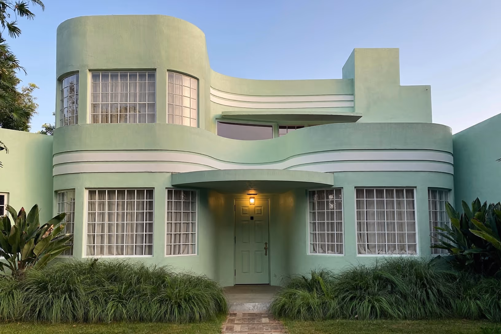 Façade d'une maison Art déco à deux niveaux enduite de crépi vert menthe, aux volumes cylindriques et bandeaux blancs horizontaux, entourée d'une végétation tropicale dense, photographiée au crépuscule.