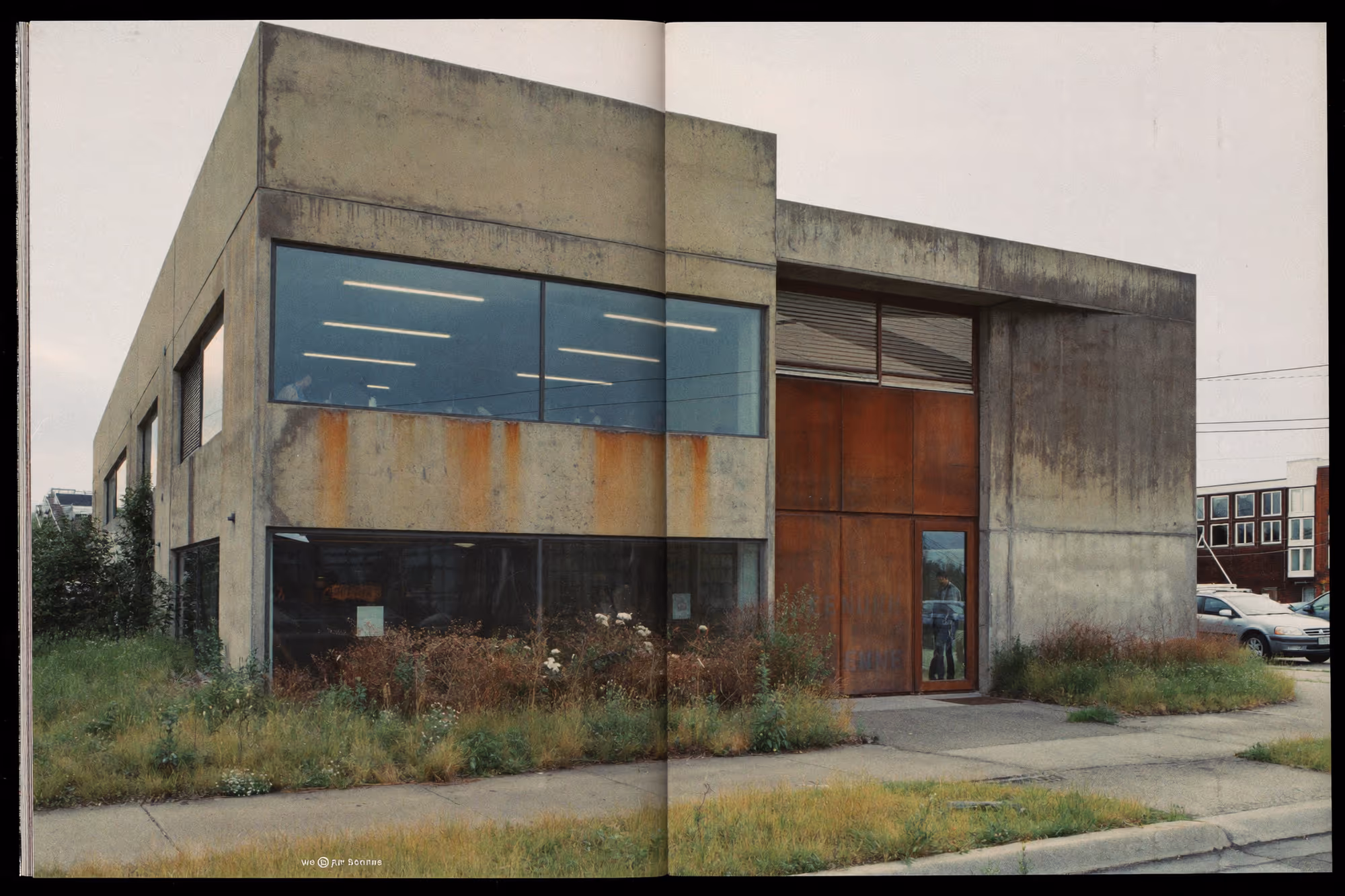 Bâtiment industriel à deux niveaux en béton brut avec grandes baies vitrées, grande porte en acier corten rouillé, végétation spontanée au pied de la façade, ciel couvert, contexte urbain périphérique.