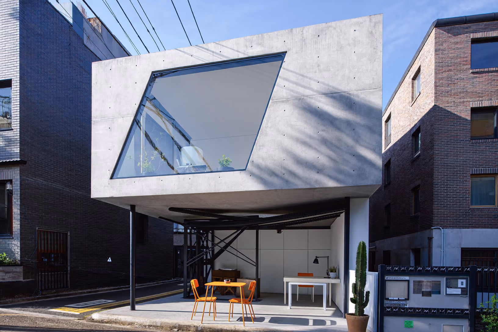 Volume cubique en béton apparent élevé sur piliers métalliques noirs, avec une large baie vitrée en trapèze asymétrique en façade ; rez-de-chaussée ouvert abritant tables et chaises orange, dans une ruelle urbaine dense.