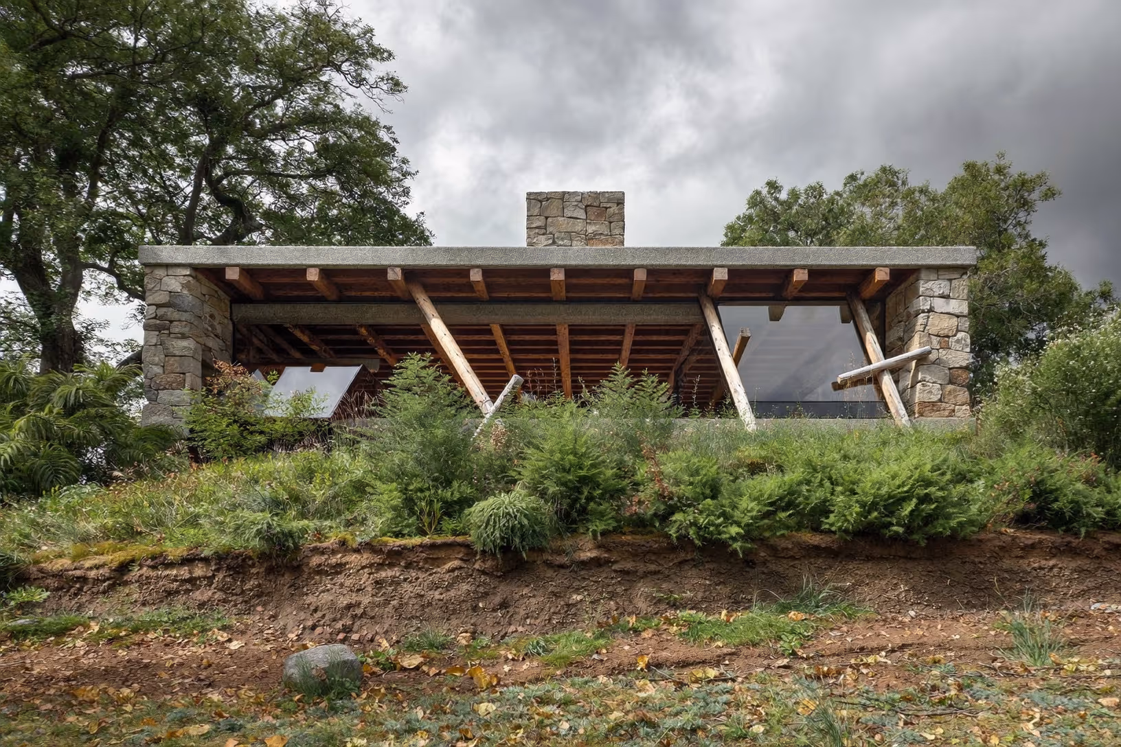Maison basse à toit-terrasse en béton, portée par des piliers de pierre appareillée brute, avec charpente en bois massif et contreventements obliques, insérée dans une pente herbeuse et arborée sous un ciel couvert.