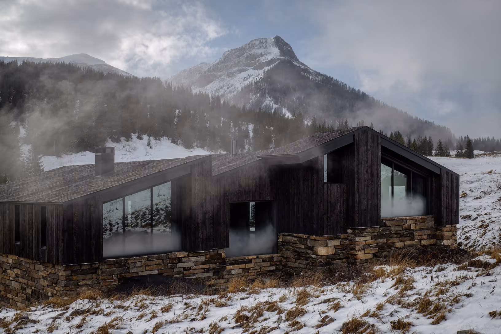Résidence de montagne à deux volumes en bois carbonisé noir sur socle de pierres appareillées, entourée de neige et de brouillard, avec un sommet alpin boisé en arrière-plan.