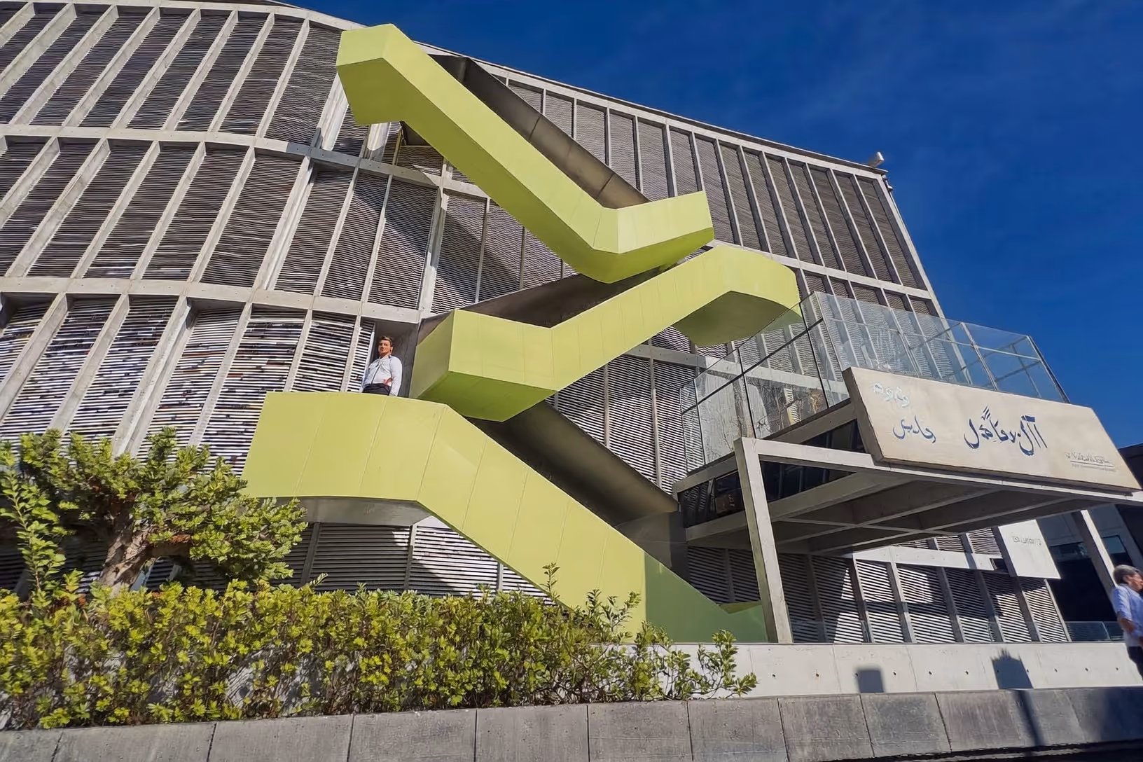 Façade d'un bâtiment en béton à lames horizontales, avec un escalier extérieur en zigzag peint en vert chartreuse, végétation arbustive au premier plan, ciel bleu vif.