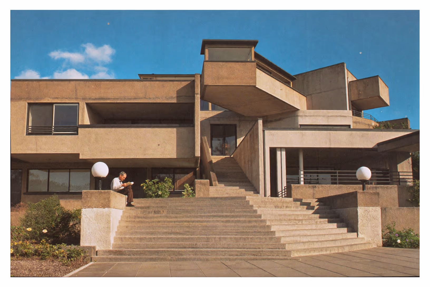 Façade d'un bâtiment brutaliste en béton clair, composée de volumes géométriques superposés et décalés, avec un large escalier central, des balcons en porte-à-faux, des globes lumineux sphériques et un homme assis lisant sur les marches.