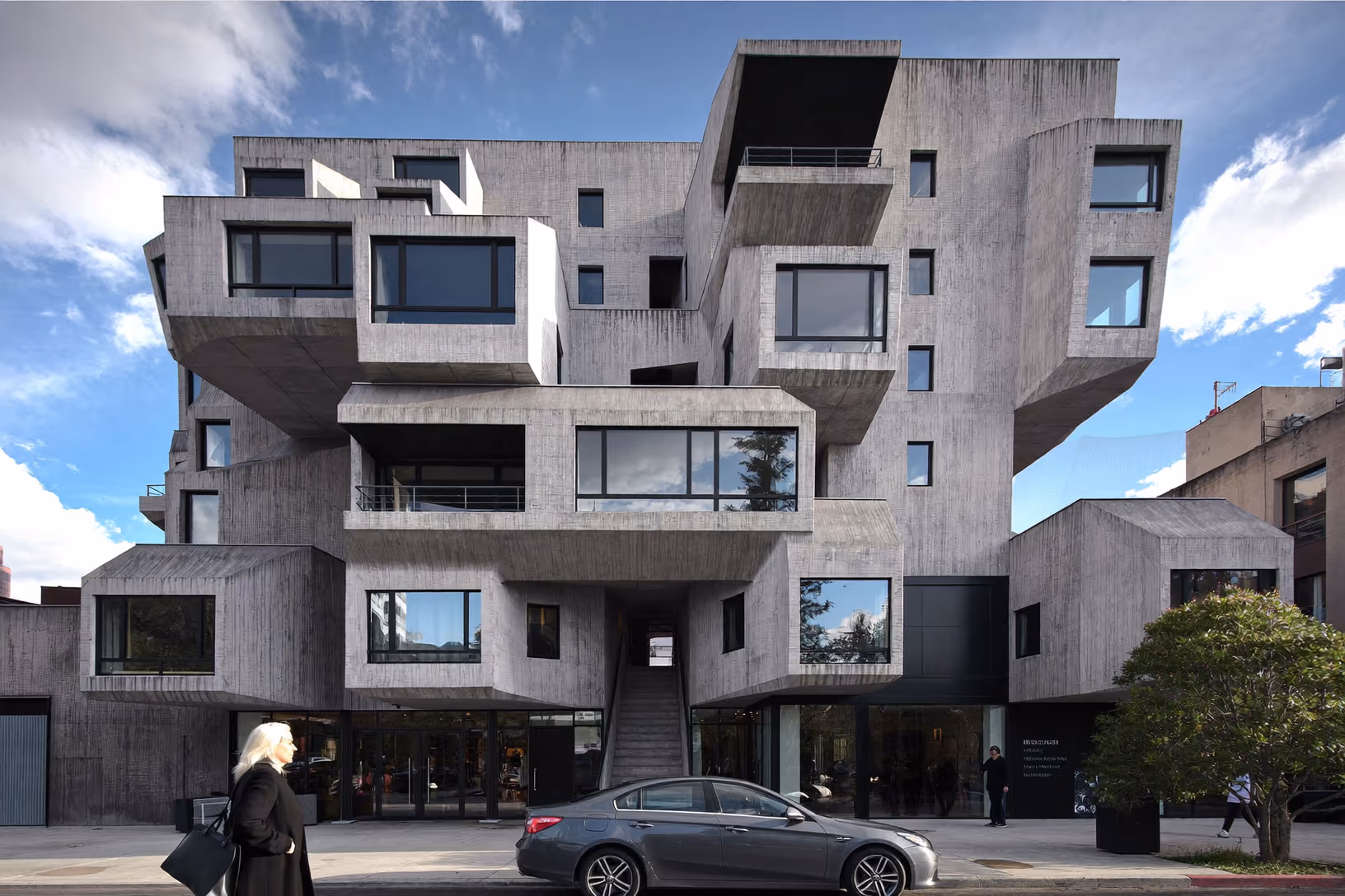 Immeuble urbain de cinq niveaux en béton brut avec volumes en porte-à-faux aux angles biseautés, fenêtres à cadres métalliques sombres, escalier extérieur central, passants et voiture au premier plan.