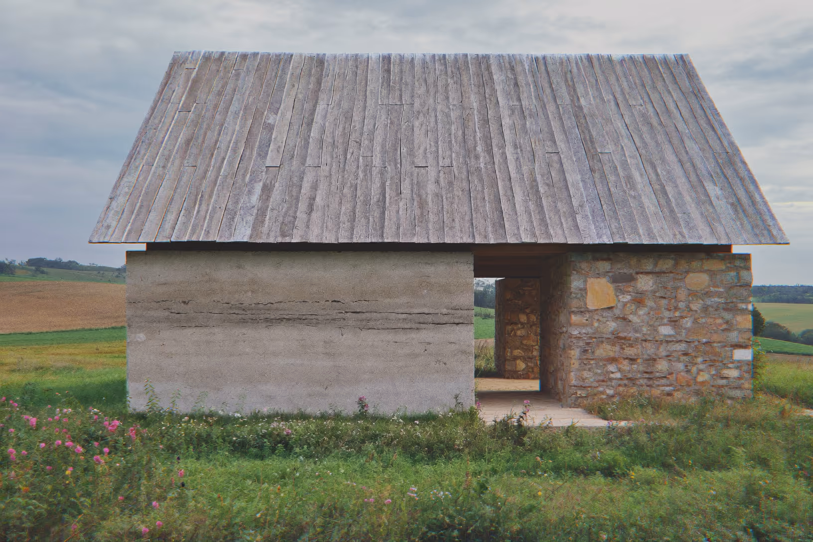 Petite construction rurale en pisé et pierre de taille, toiture à deux pans en planches de bois grisées, passage central ouvert, entourée de végétation sauvage et de champs agricoles sous un ciel couvert.