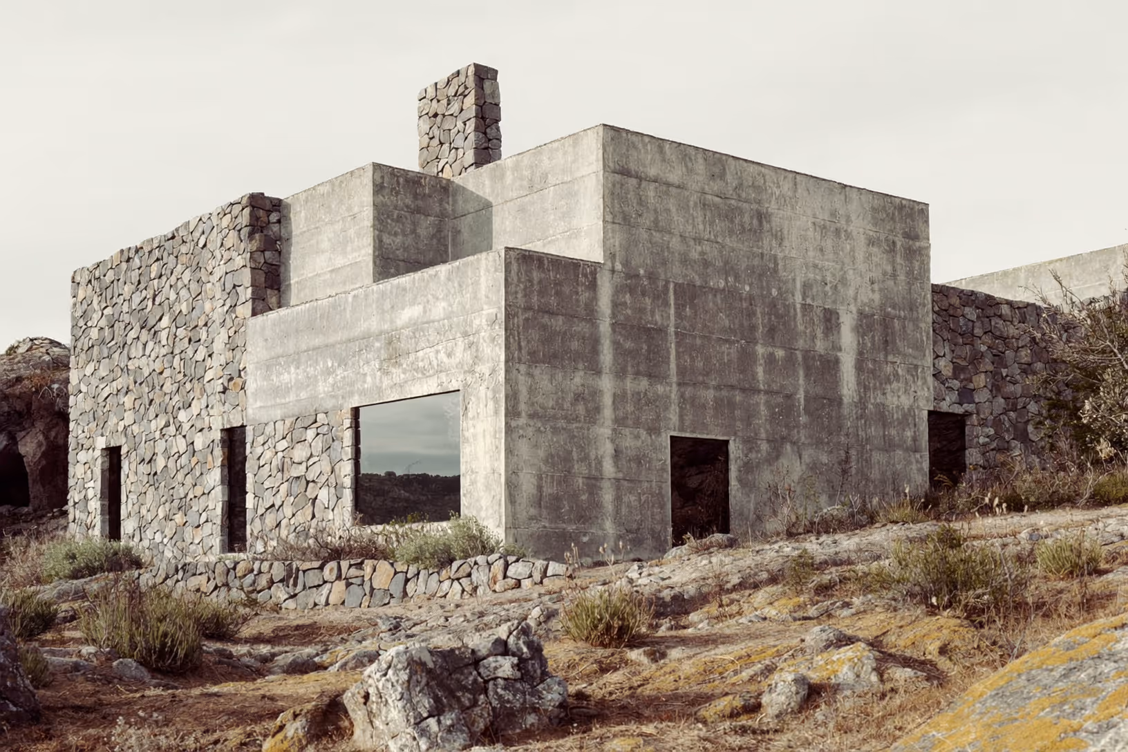 Volume architectural composé de béton brut et de murs en pierre sèche posé sur un sol rocailleux et aride, avec une cheminée en pierre en saillie sur le toit plat, ciel blanc uniforme.