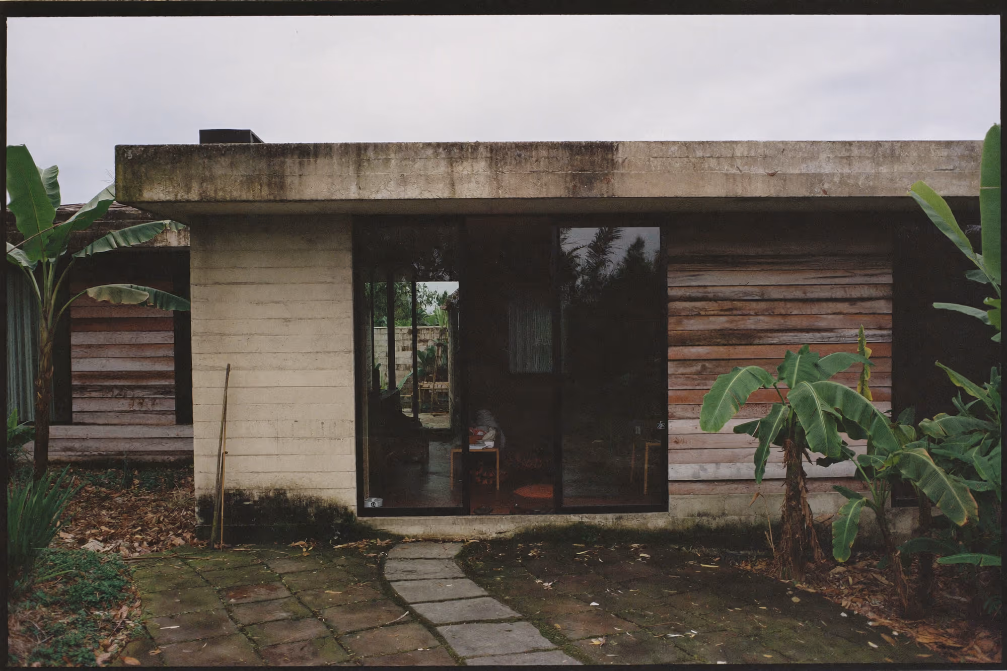 Maison de plain-pied en béton brut avec panneaux de bois aux teintes variées, large baie vitrée centrale reflétant la végétation, entourée de bananiers, allée de dalles en pierre, ciel gris uniforme.
