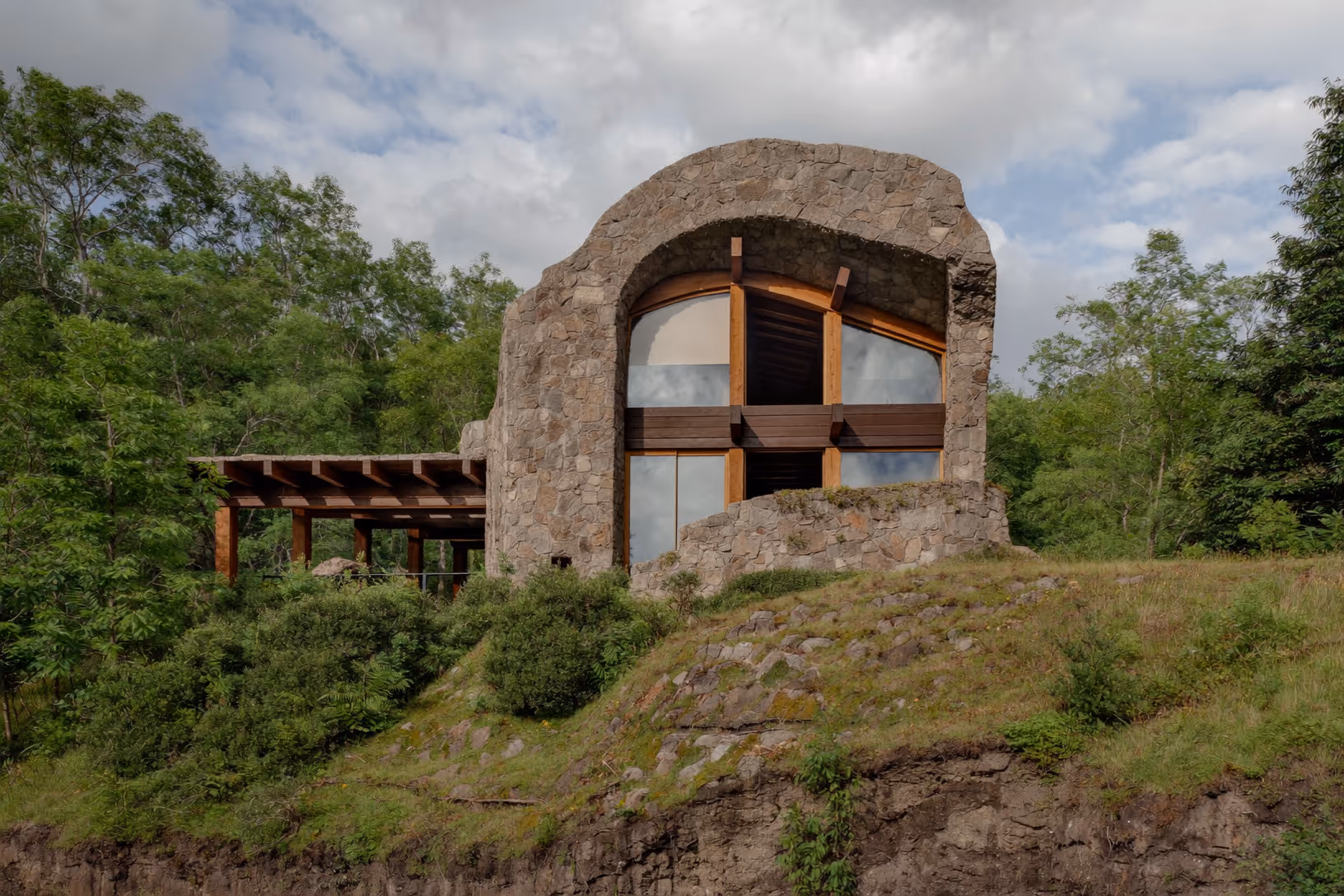 Bâtiment en pierre brute à voûte en plein cintre avec grandes baies vitrées à ossature bois, posé sur un éperon rocheux entouré de végétation verte dense, ciel nuageux en arrière-plan.