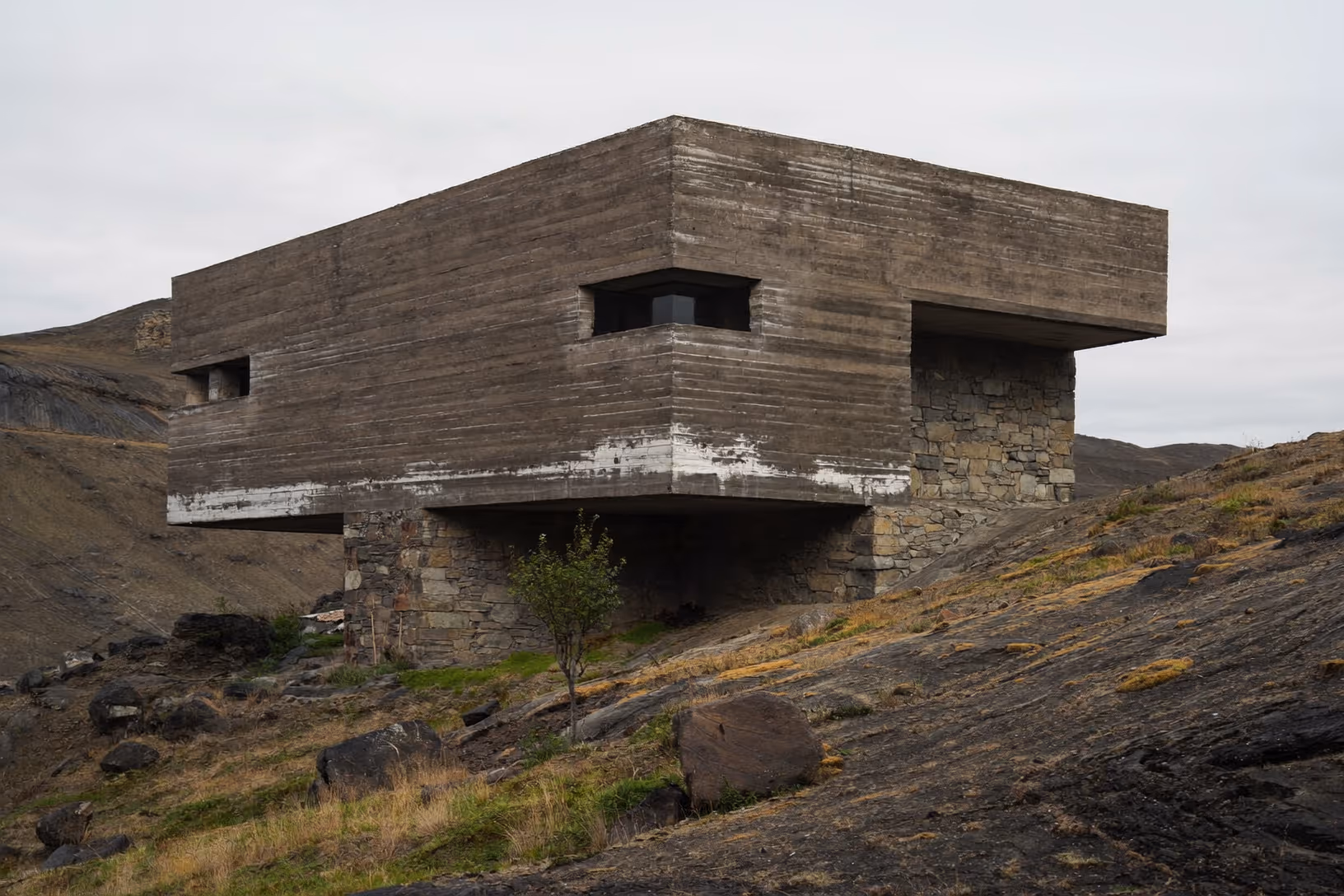 Volume parallélépipédique en béton strié posé en porte-à-faux sur deux piliers de pierre sèche, implanté sur une pente rocheuse aride sous un ciel gris uniforme ; un arbre isolé pousse au pied de la structure.