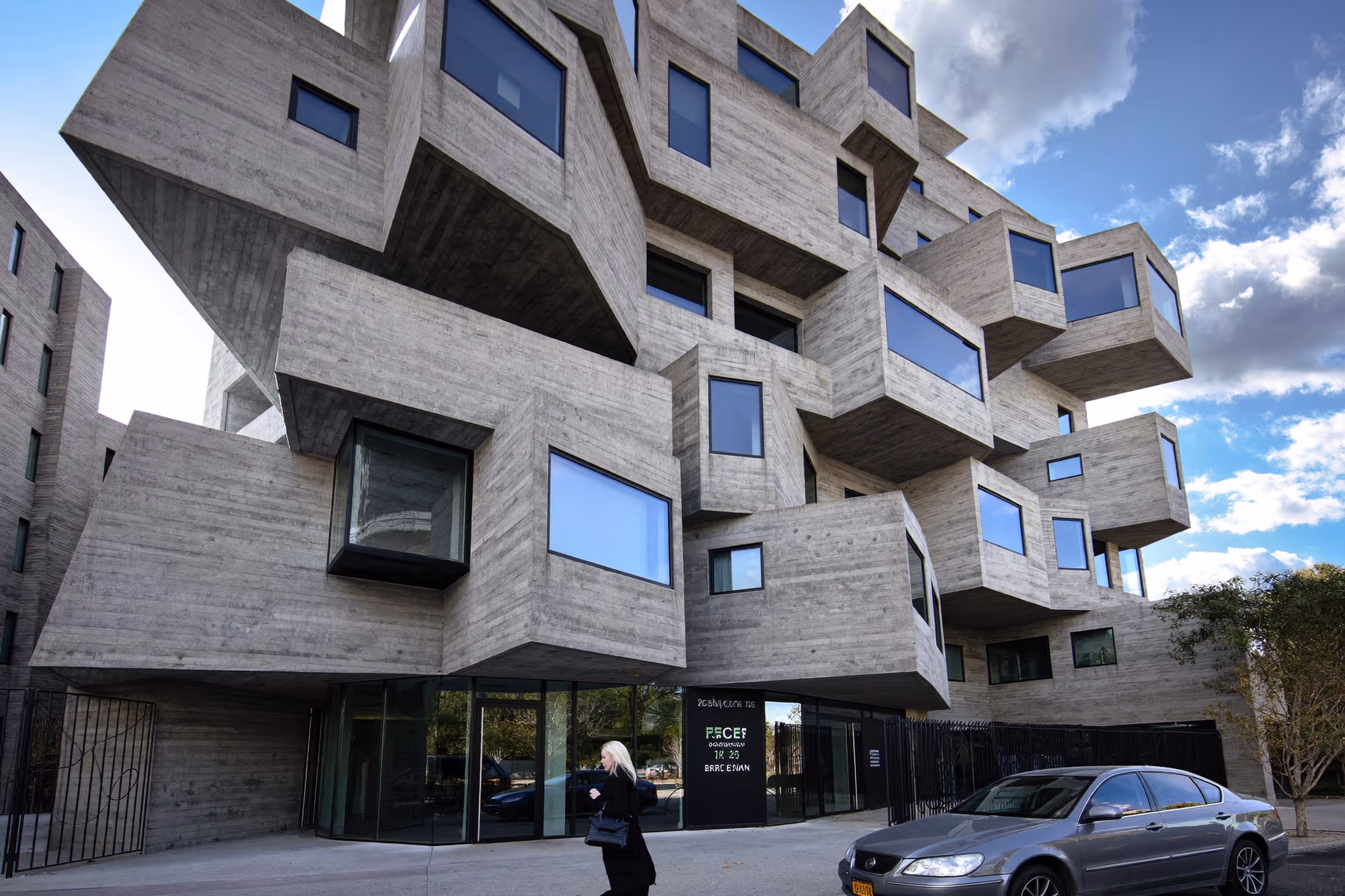 Immeuble collectif en béton brut à volumes cubiques asymétriques en saillie et en retrait sur plusieurs niveaux, avec fenêtres rectangulaires sombres, photographié depuis la rue avec une passante et une voiture au premier plan.