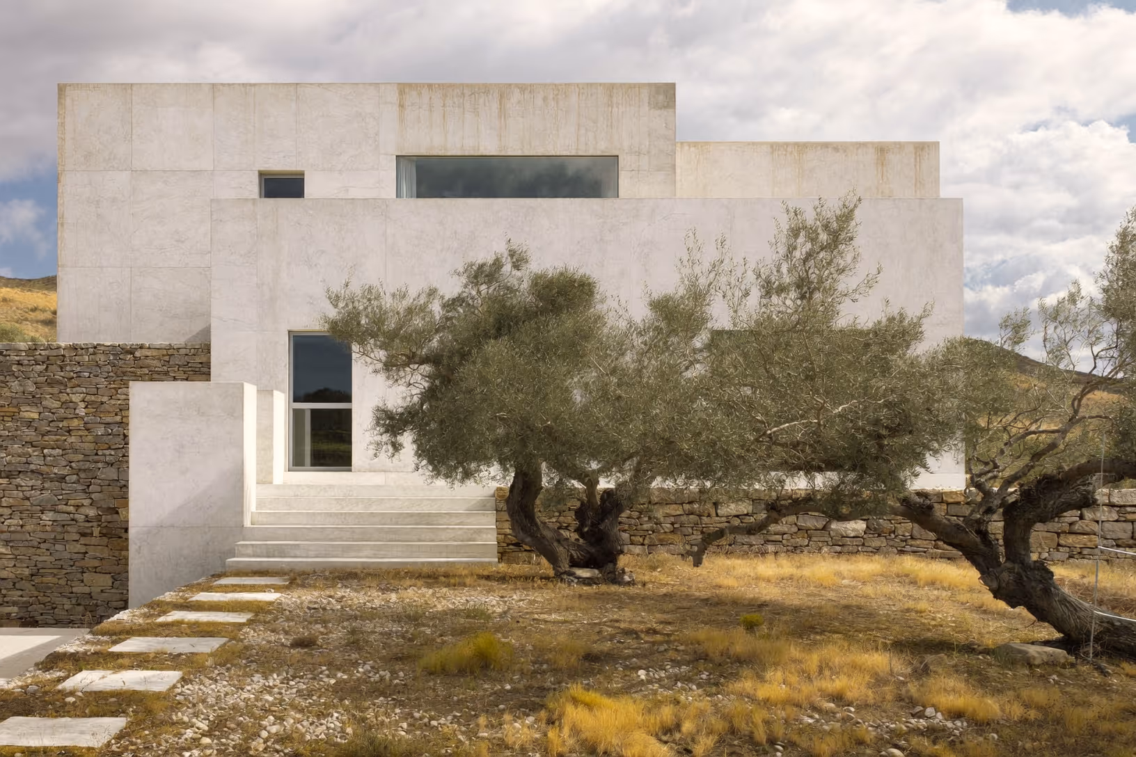 Façade d'une maison en béton apparent aux volumes orthogonaux empilés, avec un escalier central et deux oliviers anciens au premier plan sur un terrain aride, mur de pierre sèche à gauche.