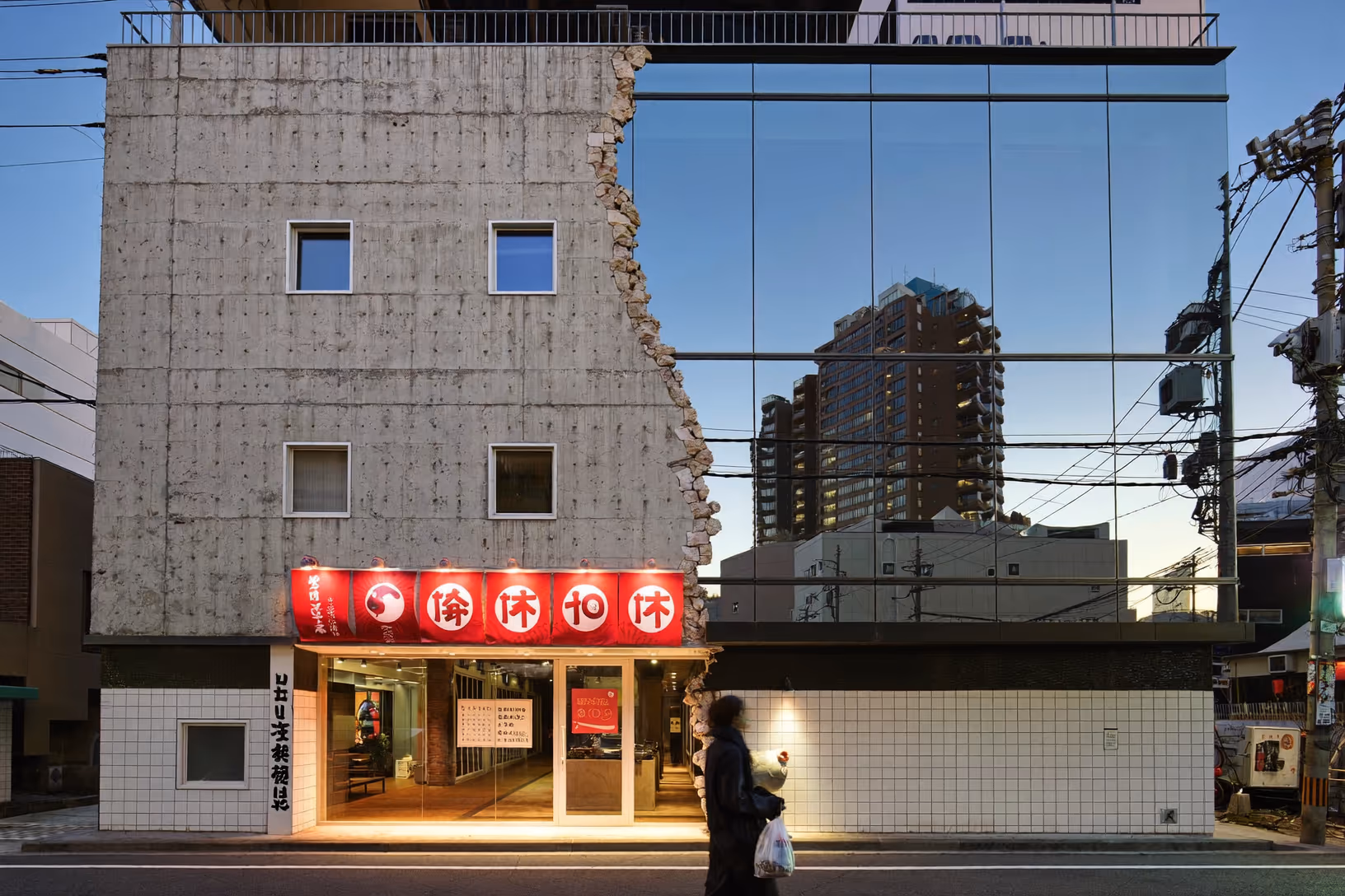 Façade d'un immeuble urbain en contexte asiatique dense : à gauche un mur de béton brut aux joints de coffrage apparents, dont l'arête droite s'effiloche en brique exposée irrégulière ; à droite une façade entièrement vitrée miroir reflétant les immeubles alentour et le ciel bleu du soir. Au rez-de-chaussée, une enseigne lumineuse rouge à caractères japonais surmonte l'entrée carrelée de blanc. Un passant traverse au premier plan.