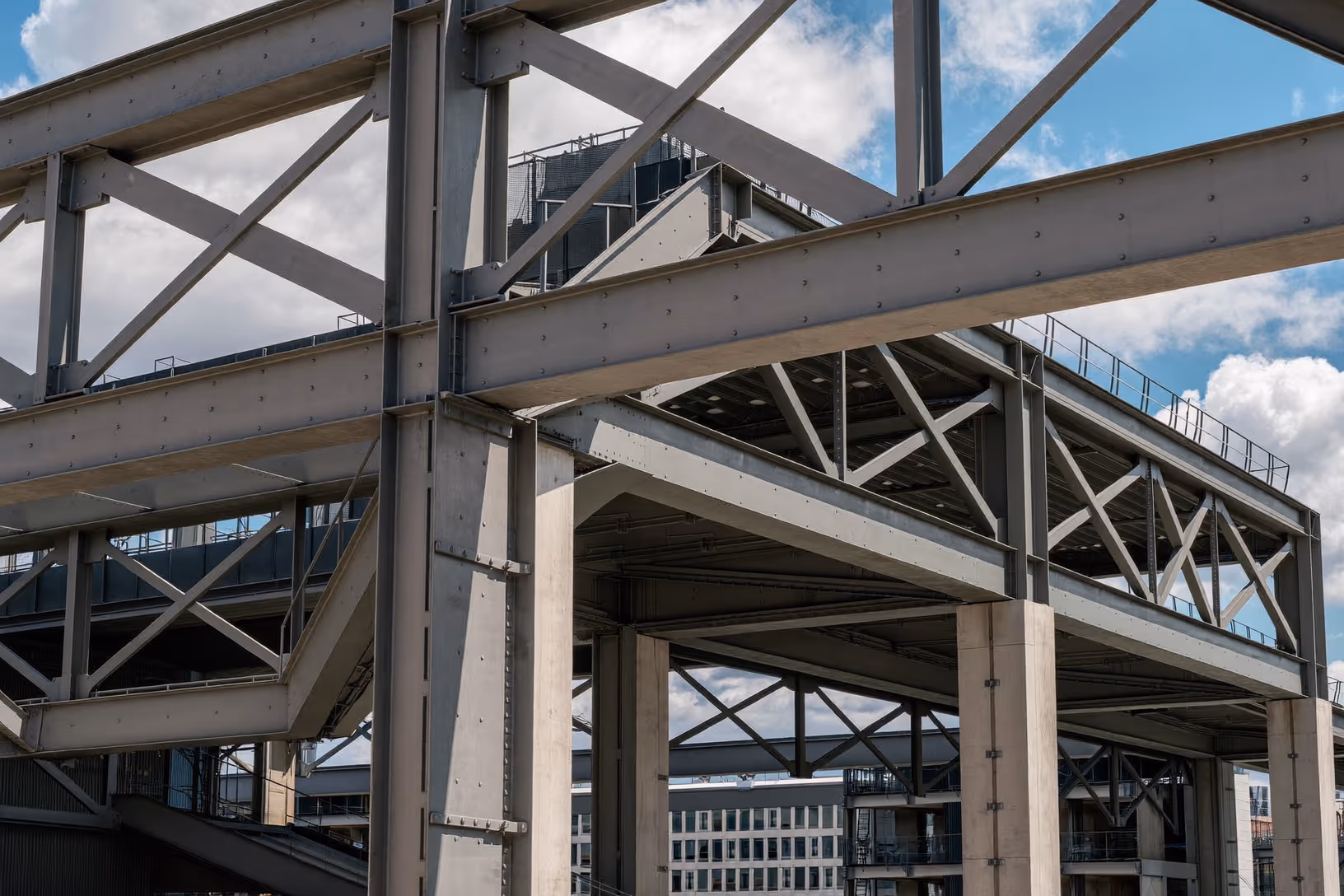 Poutres en treillis d'acier gris photographiées en contre-plongée, avec colonnes boulonnées et tissu urbain visible en arrière-plan sous un ciel nuageux.