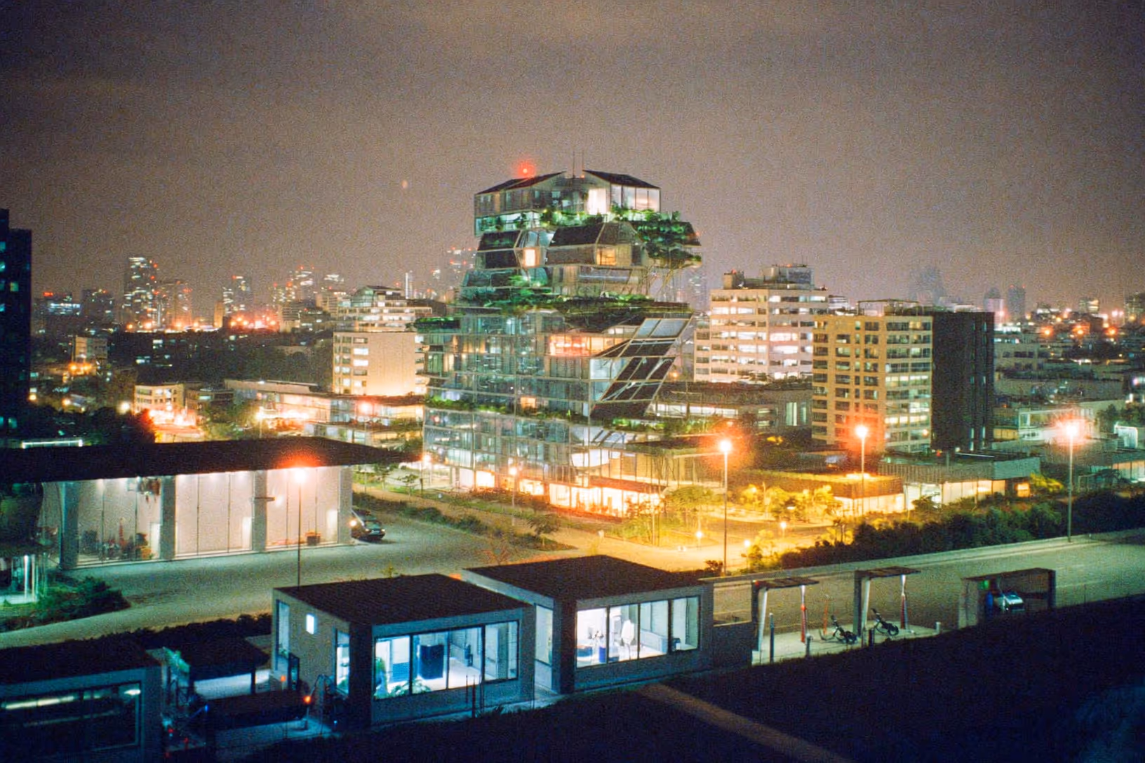 Tour de verre et d'acier photographiée de nuit, avec des terrasses étagées chargées de végétation, dans un tissu urbain dense éclairé par les lumières de la ville.