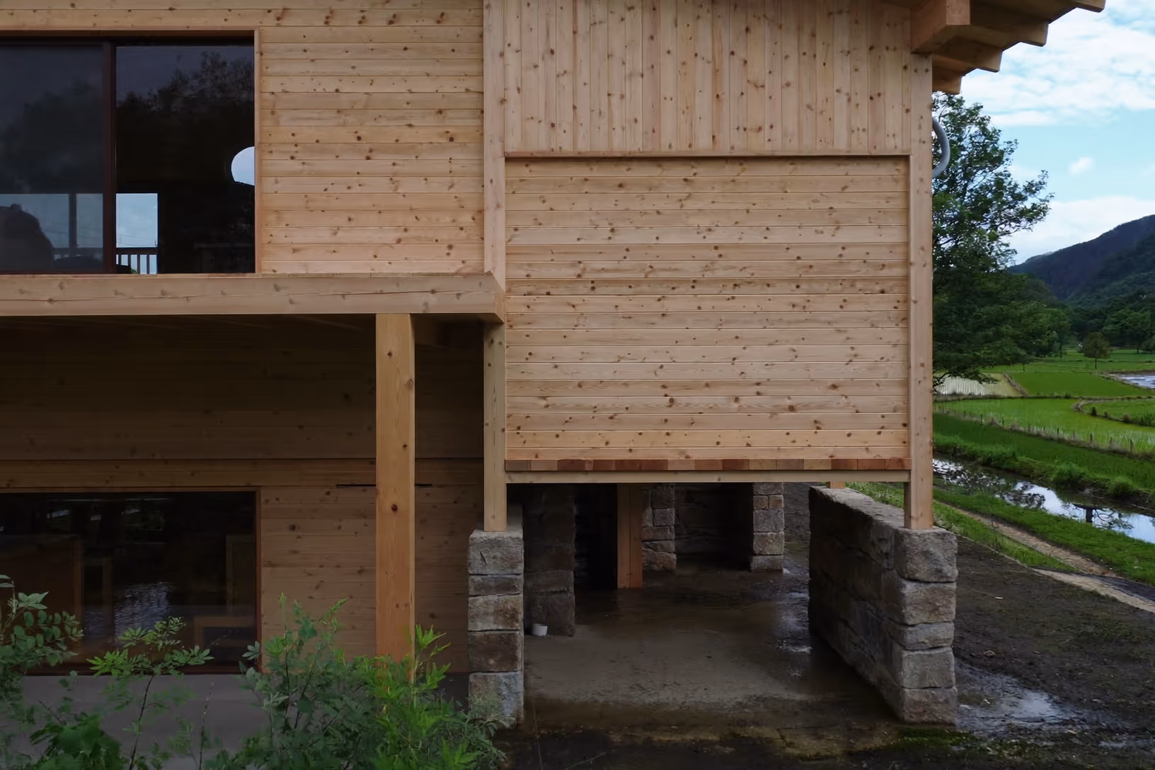 Façade en bardage bois clair d'une maison élevée sur des piliers en pierre taillée, avec en arrière-plan une vallée verdoyante, des rizières et un cours d'eau.