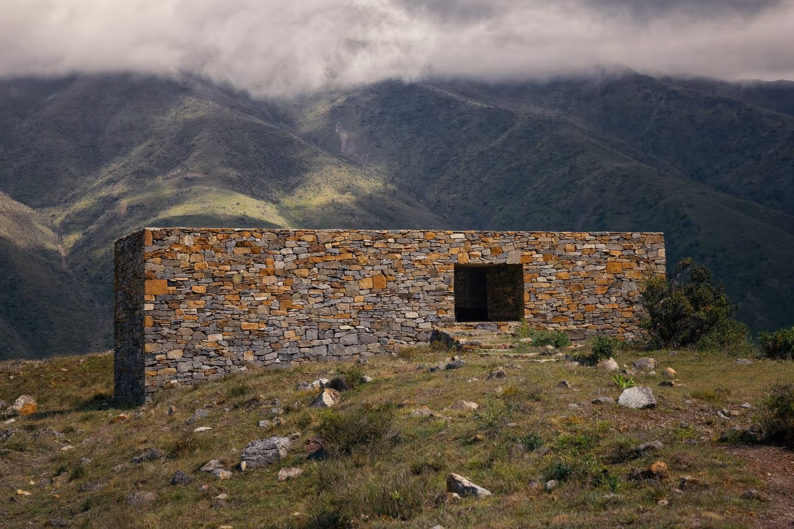 Volume parallélépipédique bas en pierre sèche appareillée, seul sur un promontoire herbeux et rocheux, avec un unique percement rectangulaire en façade, devant une vallée montagneuse aux versants verts sous un ciel nuageux.