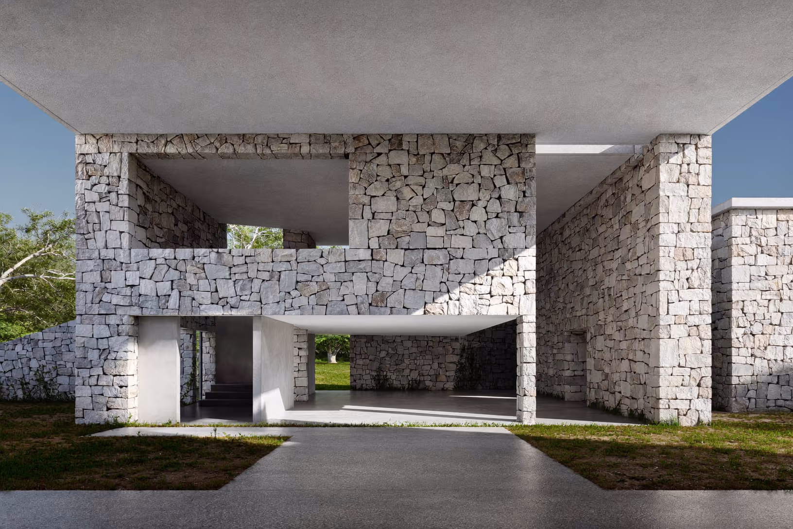 Façade frontale d'une maison à deux niveaux composée de murs en pierres calcaires irrégulières et de dalles horizontales en béton gris, ouverte sur un espace traversant au rez-de-chaussée, entourée de végétation basse.