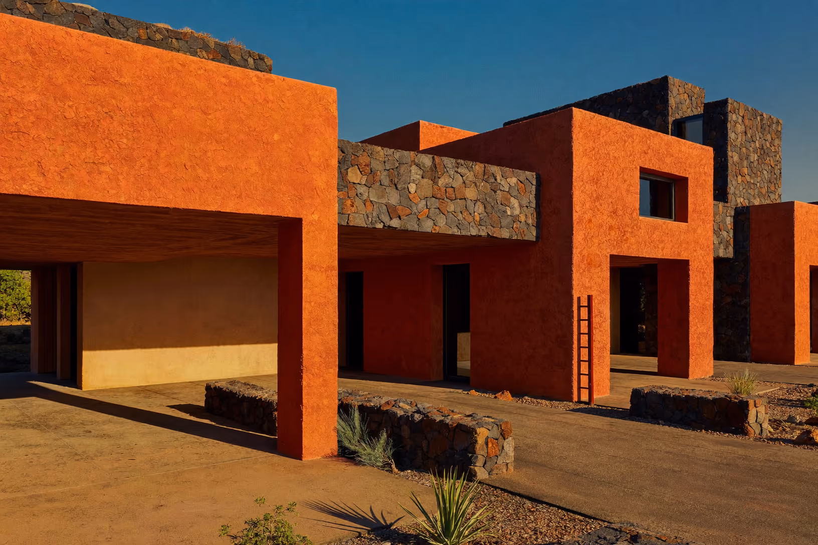 Volumes cubiques enduits de crépi ocre-rouge, avec bandeaux en pierre volcanique brute, sol de gravier minéral, quelques agaves, lumière rasante de fin de journée, ciel bleu acier.