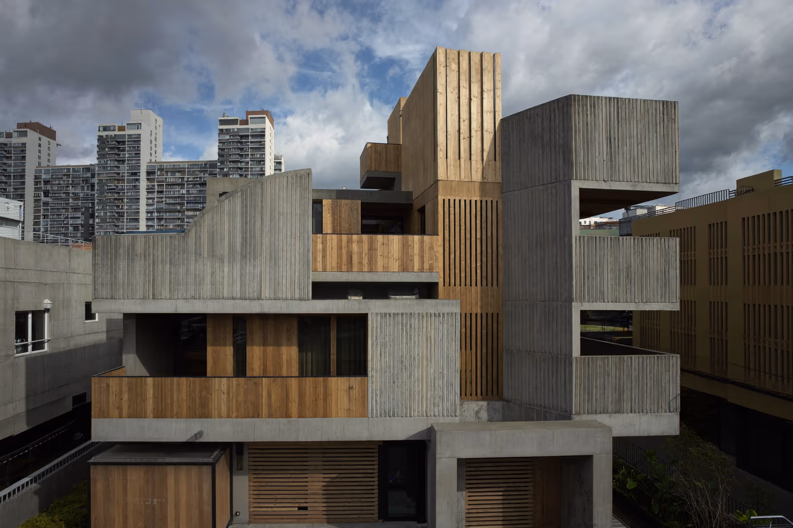 Façade d'un bâtiment urbain composé de volumes cubiques en béton strié à coffrage bois, avec inserts en lattes de bois naturel, décalés en hauteur sur fond de tours résidentielles et ciel nuageux.