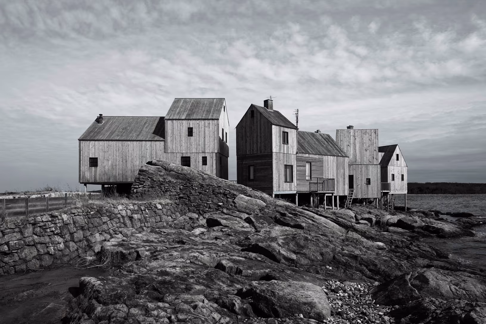 Photographie en noir et blanc d'un ensemble de petits volumes en bois à toits à deux pentes, sur pilotis, dressés sur un affleurement rocheux en bord de mer sous un ciel nuageux.