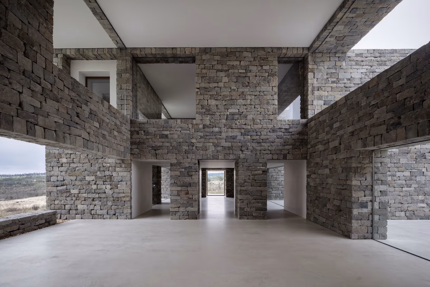 Vue intérieure d'un grand espace en pierre de taille grise sombre, avec une enfilade de portiques ouverts sur un couloir central, sol en béton poli, plafond blanc, et ouvertures latérales cadrant un paysage aride.