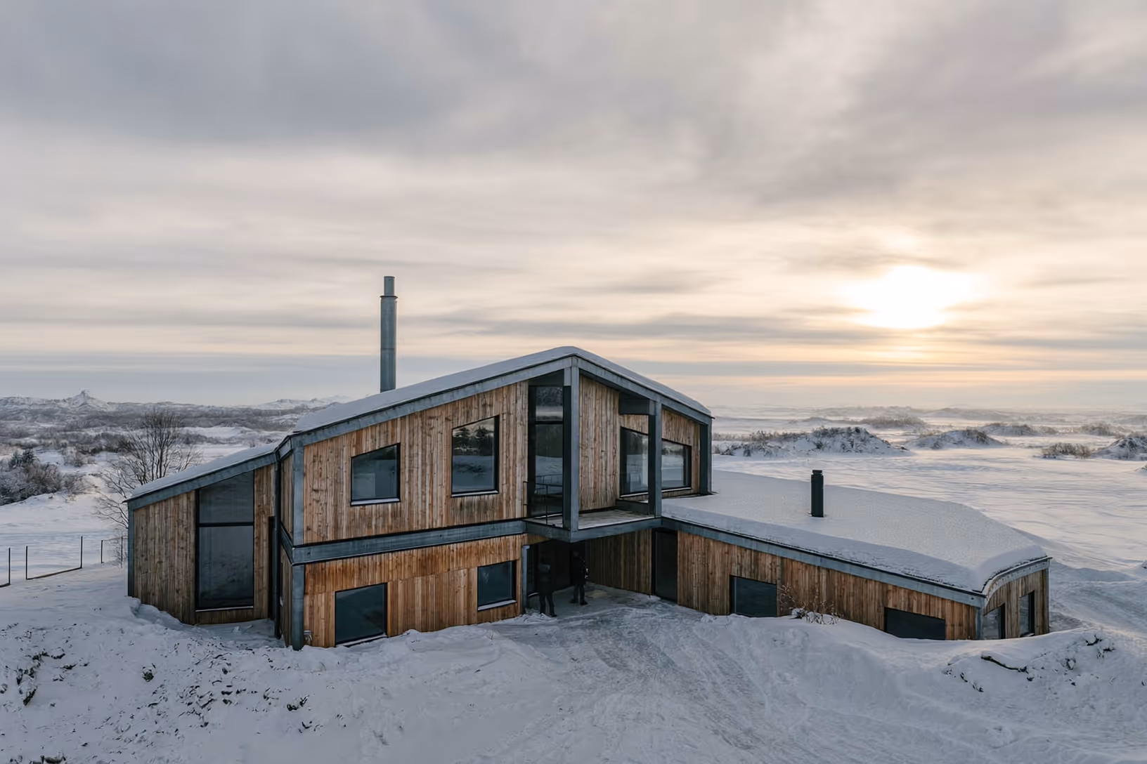 Maison en bardage de bois naturel aux volumes angulaires imbriqués, posée dans un paysage entièrement enneigé, sous un ciel nuageux traversé d'une lumière rasante ; une cheminée métallique fine s'élève au-dessus du toit.