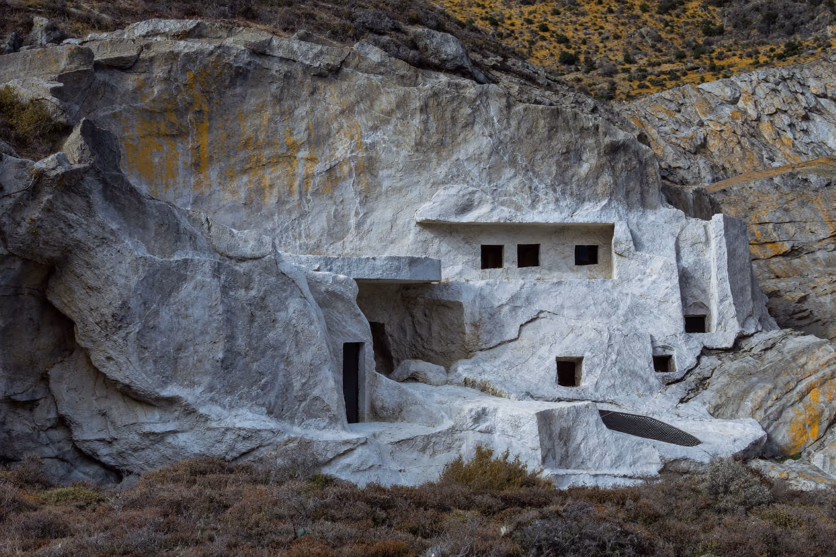 Structure habitée creusée dans une falaise de roche blanche marbréenne, avec des ouvertures rectangulaires sombres taillées dans la masse, entourée de végétation aride et broussailleuse sur un flanc de colline pierreux.