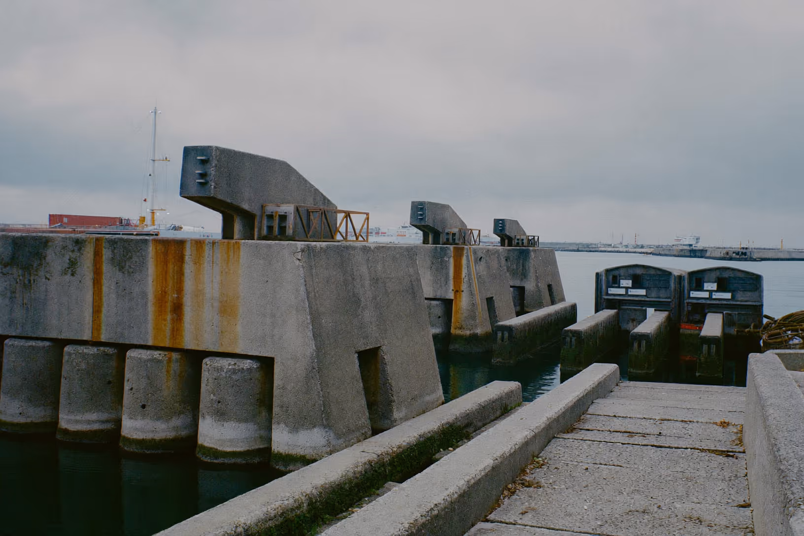 Écluses ou vannes de digue en béton gris avec piliers cylindriques, traces de rouille ocre sur les parements, eau vert-gris au premier plan, ciel couvert, port industriel en arrière-plan.
