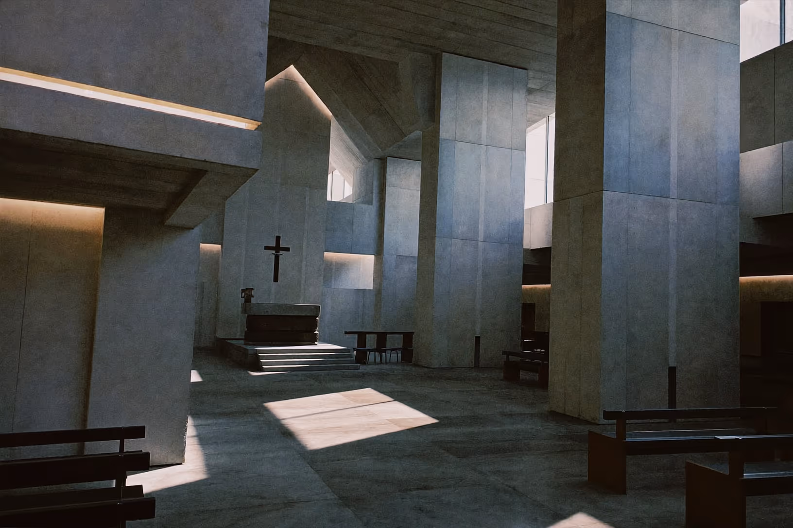 Intérieur d'une chapelle contemporaine en béton brut : hauts piliers géométriques, faisceaux de lumière naturelle projetés en diagonale sur le sol en pierre, autel surélevé et croix sombre au fond, bancs en bois foncé.