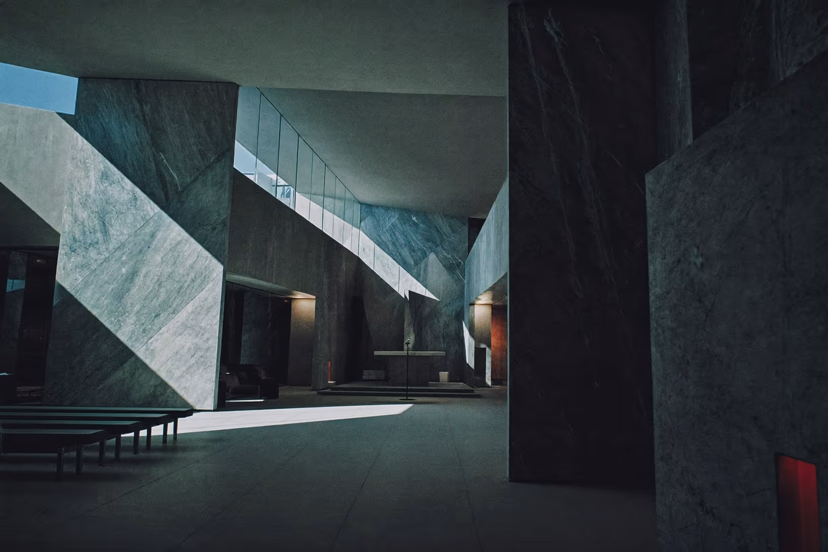 Intérieur monumental habillé de marbre sombre veiné de gris-vert, traversé par un faisceau de lumière naturelle oblique ; bancs bas à gauche, autel simplifié en fond, mezzanine vitrée en hauteur.